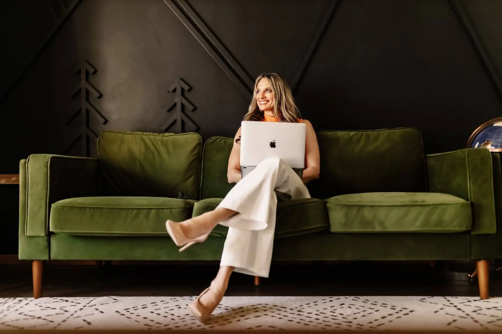 A woman with long blonde hair sitting on a green sofa, smiling, holding a laptop on her lap in a modern room with dark walls.