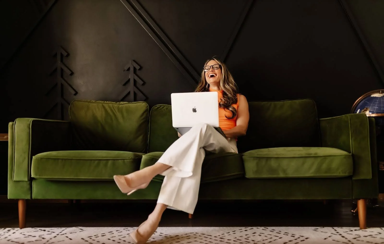 Woman sitting on a green sofa, smiling and holding a silver laptop, with a dark wall background and a blue globe on a side table.