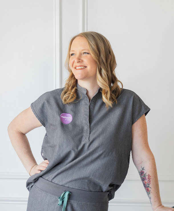 A woman with blonde hair styled in loose waves, smiling and looking off to the side. She is wearing a dark gray uniform with a purple button that says 'midwifery' and has a floral tattoo on her left forearm. Her right hand rests on her hip.