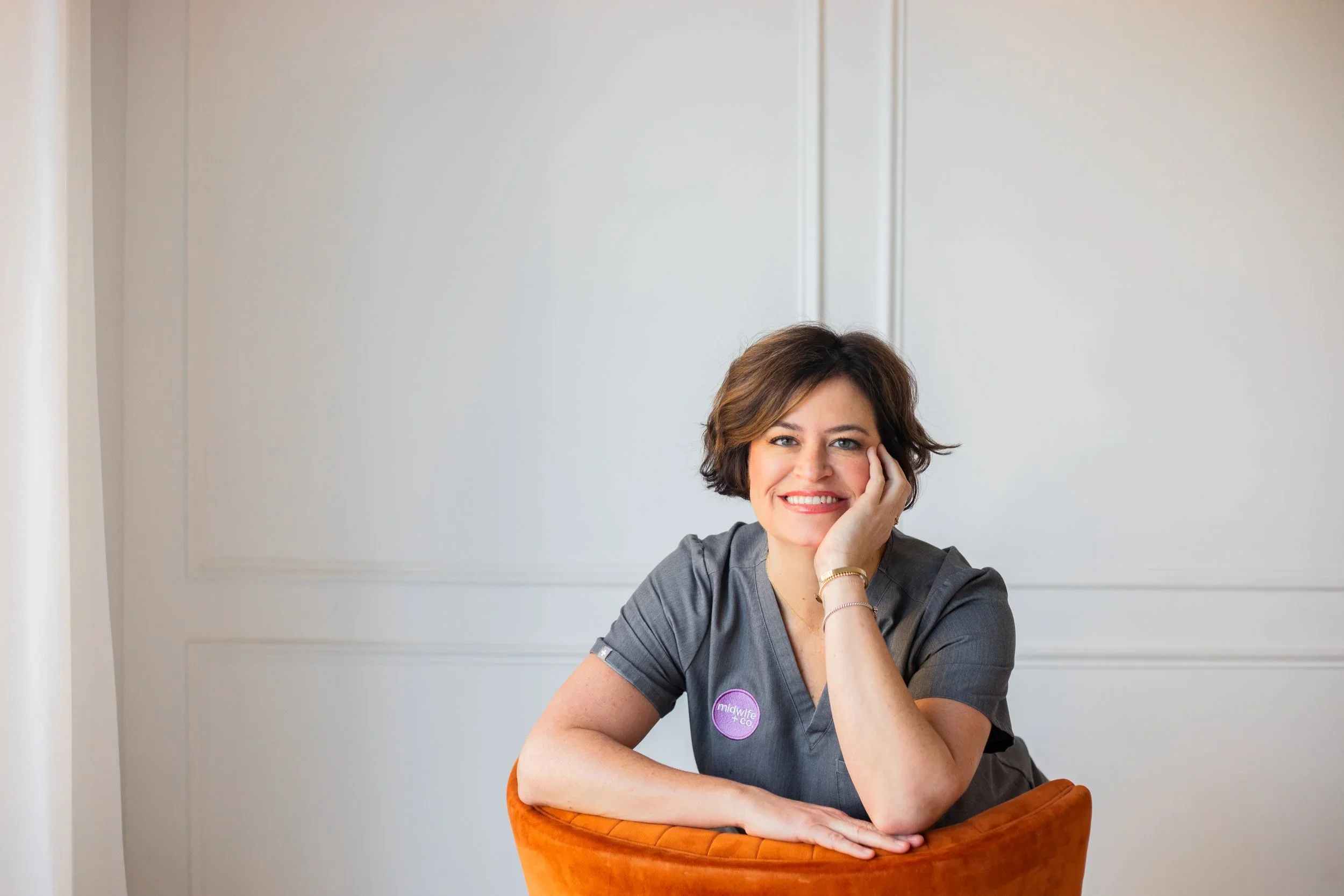 A smiling woman with short brown hair and wearing gray scrubs sitting at a table with an orange chair, resting her head on her hand, in front of a plain light-colored wall.
