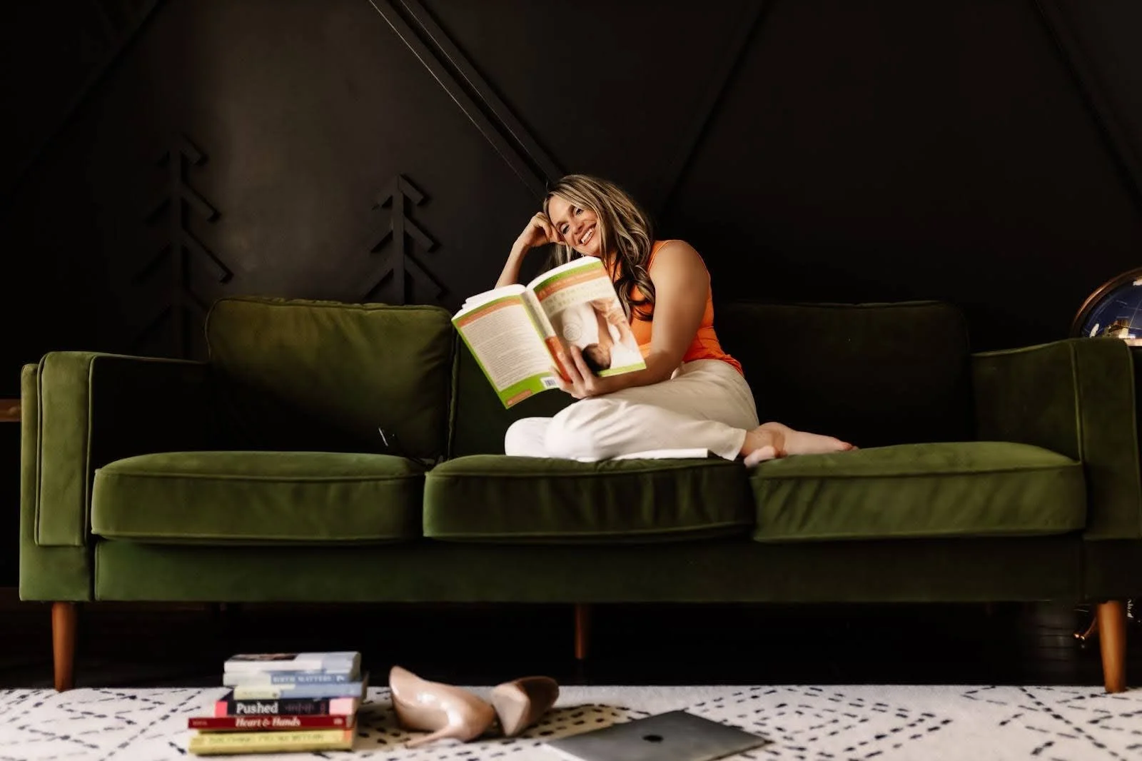 A woman sitting on a dark green sofa, reading a book, wearing an orange top and white pants, smiling, in a cozy room with dark walls.