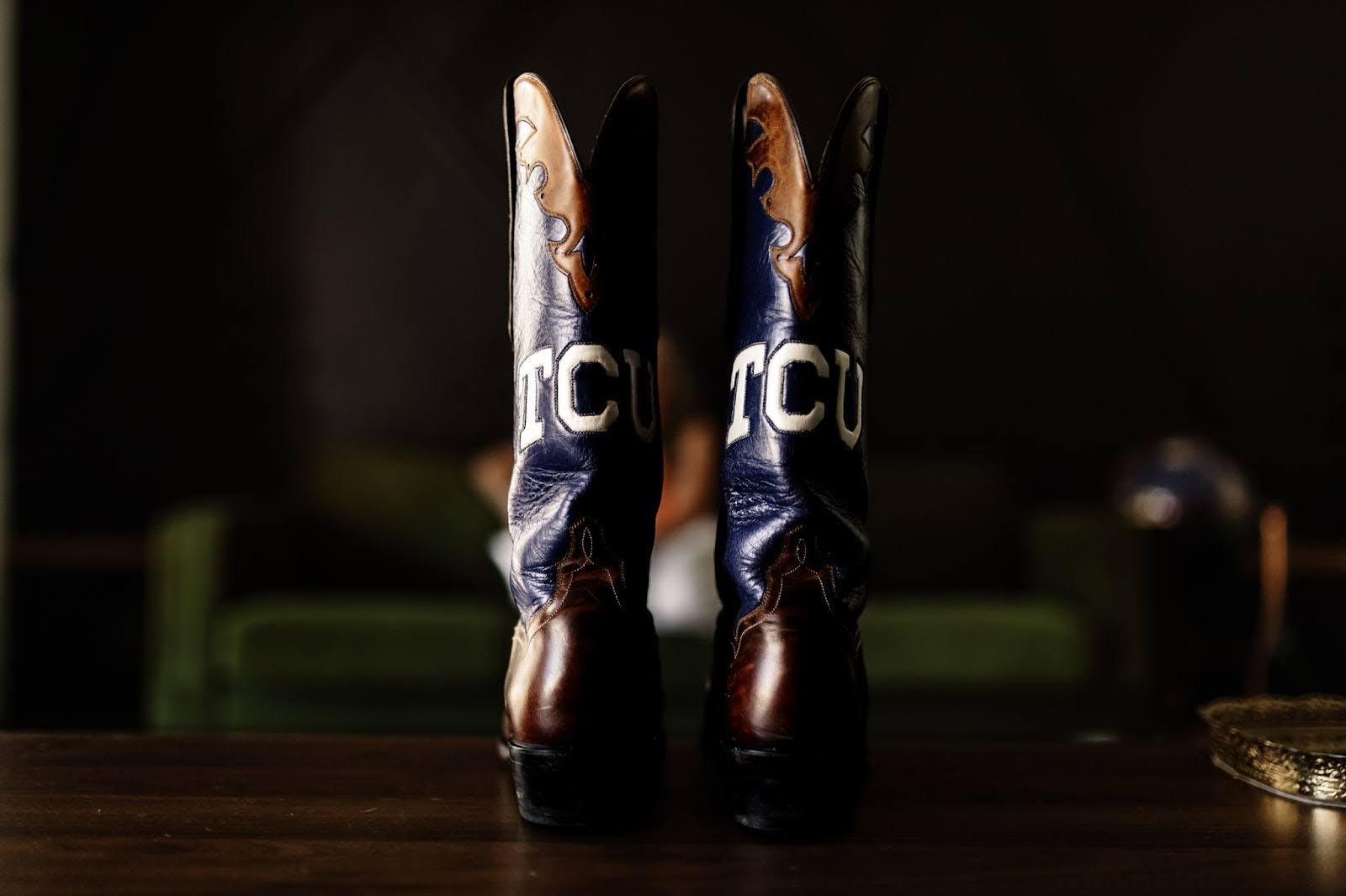 A pair of cowboy boots with the letters 'TCU' on them, placed on a wooden surface. The boots are mostly dark with some light tan and blue details.