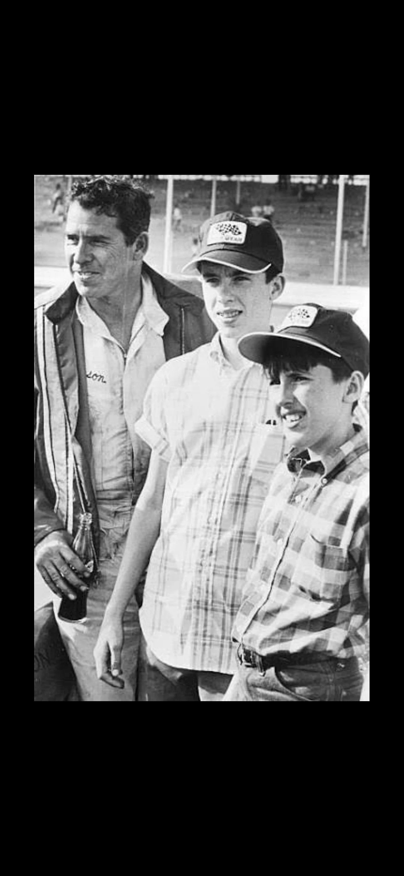 Black and white photo of three young men and an older man standing outdoors, possibly at a racing event, with a grassy field and fence in the background. The young men are wearing caps, and the older man is holding a drink.