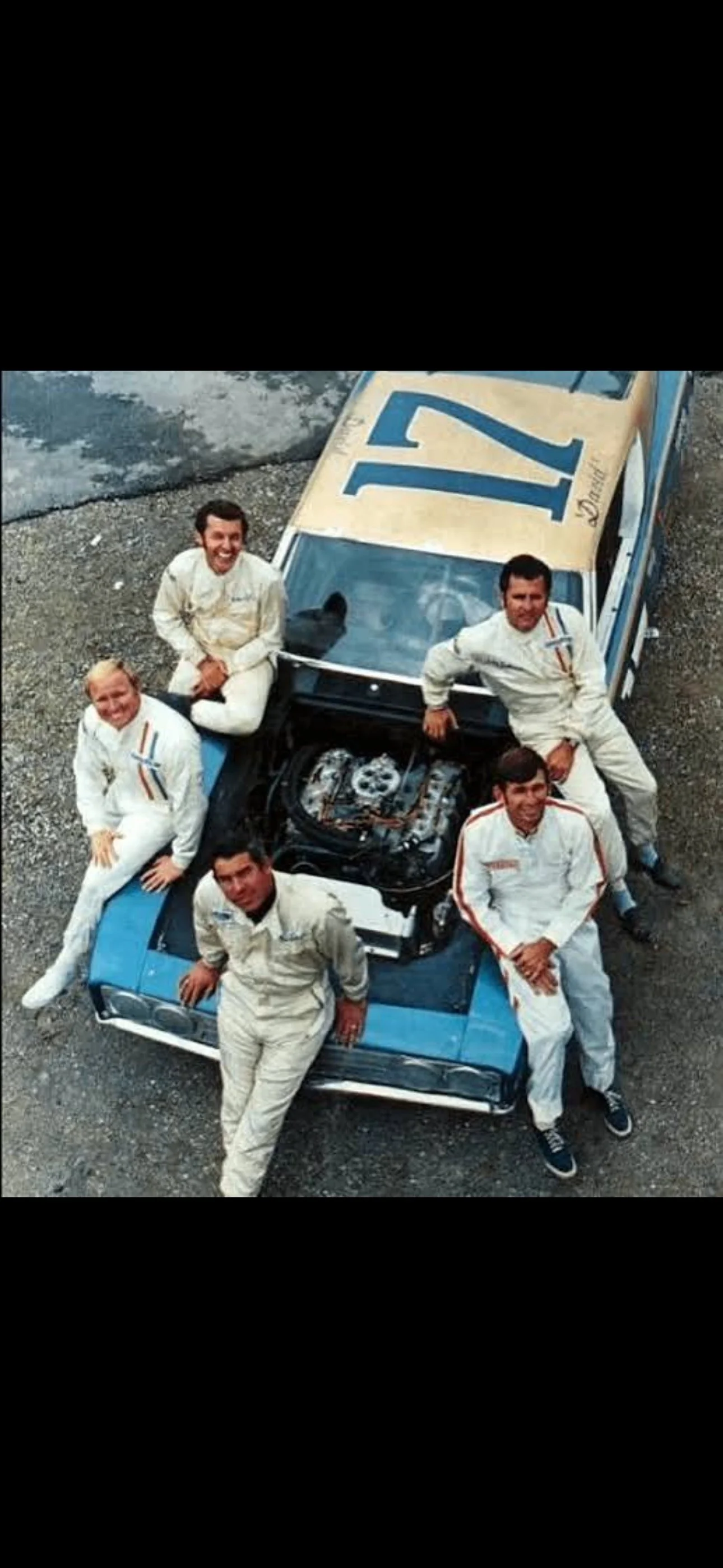 The image shows five men in racing suits posing around a race car with the hood open, revealing the engine, on a gravel surface. The car has the number 21 on the roof.