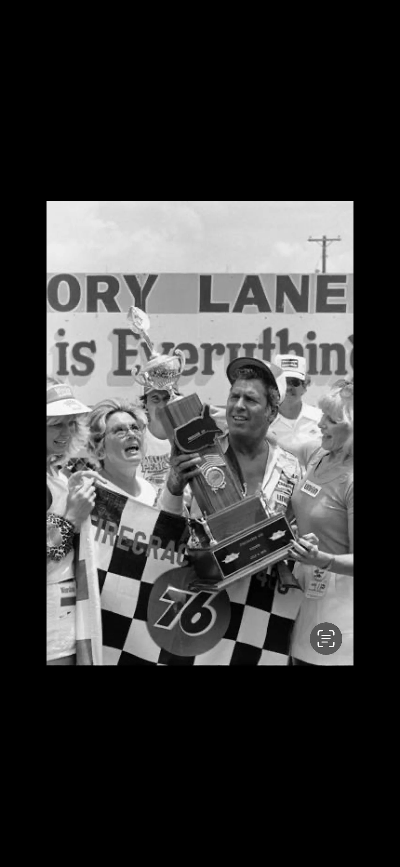 A black and white photo of a victorious NASCAR driver holding a trophy, surrounded by cheering people, at a racing event with a sign reading 'History Lane is Everything' in the background.