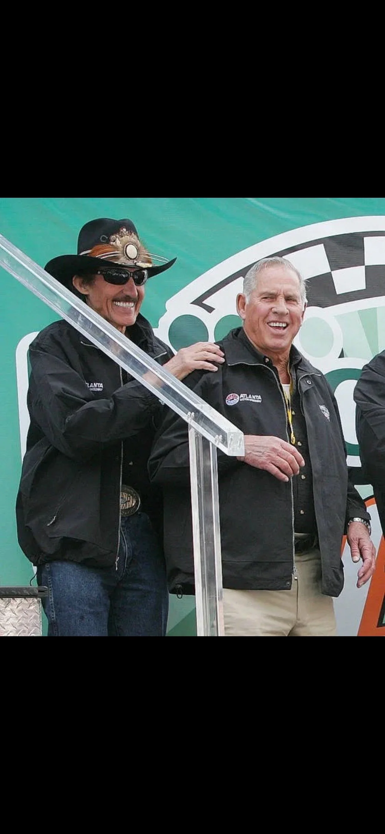 Two men standing outdoors, one wearing sunglasses and a hat with decorations and the other smiling, both dressed in black jackets, in front of a green backdrop with a logo featuring a checkered flag.
