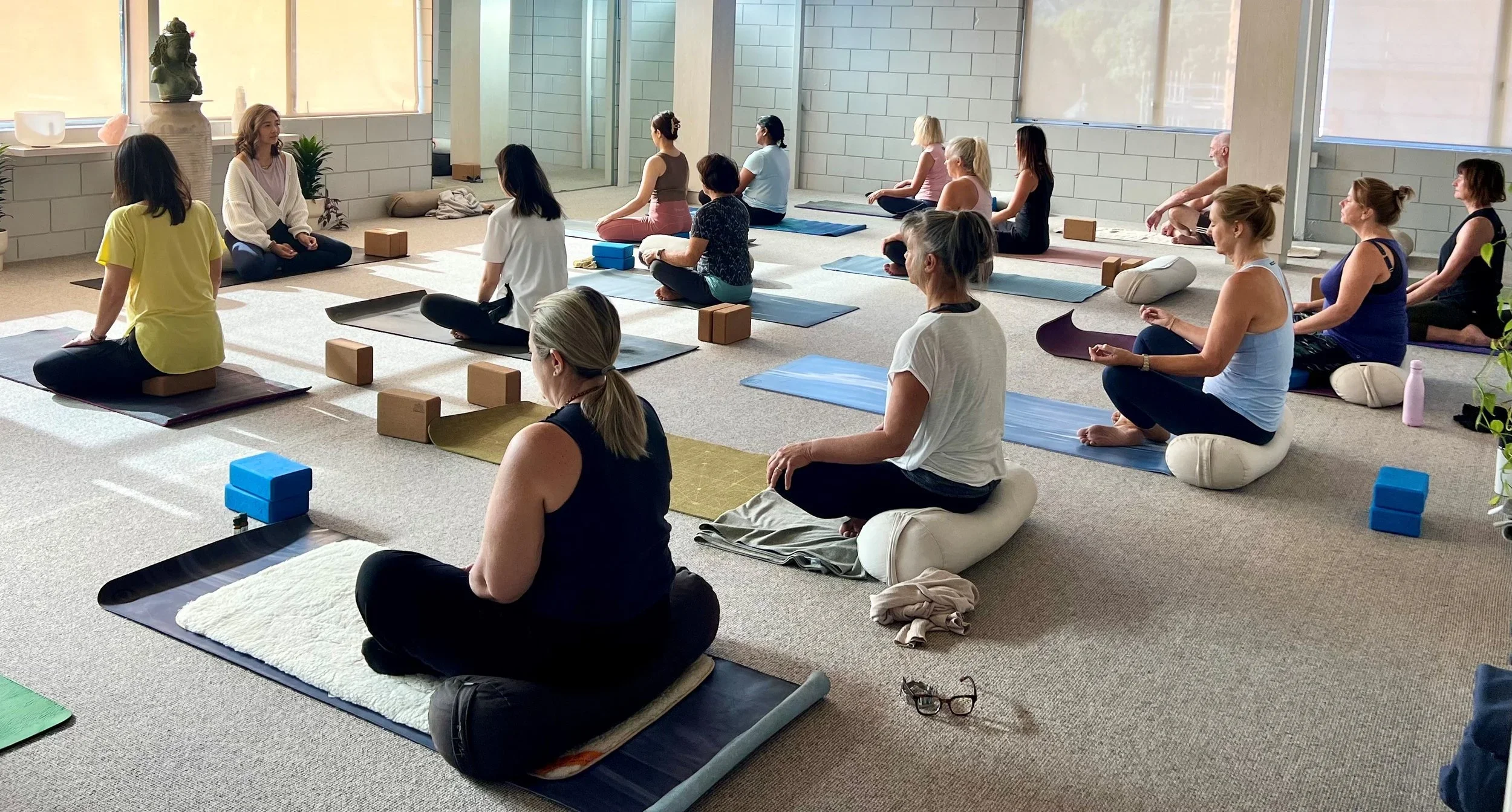 Group of people practicing yoga or meditation in a bright studio, seated on mats with blocks and cushions, facing a woman leading the session.