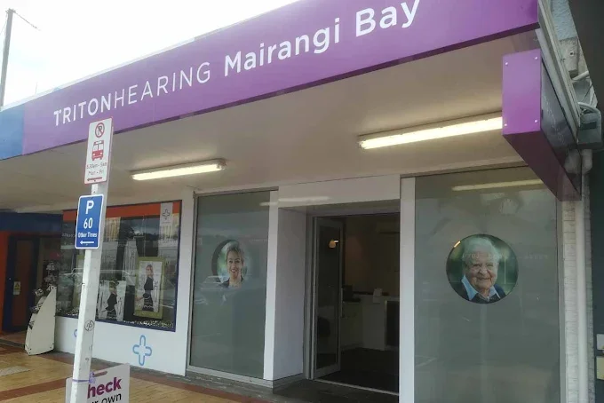The exterior of a clinic named TritoHearing Mairangi Bay with a purple sign, large front windows displaying portraits of elderly people, parking signs, and part of a sidewalk.