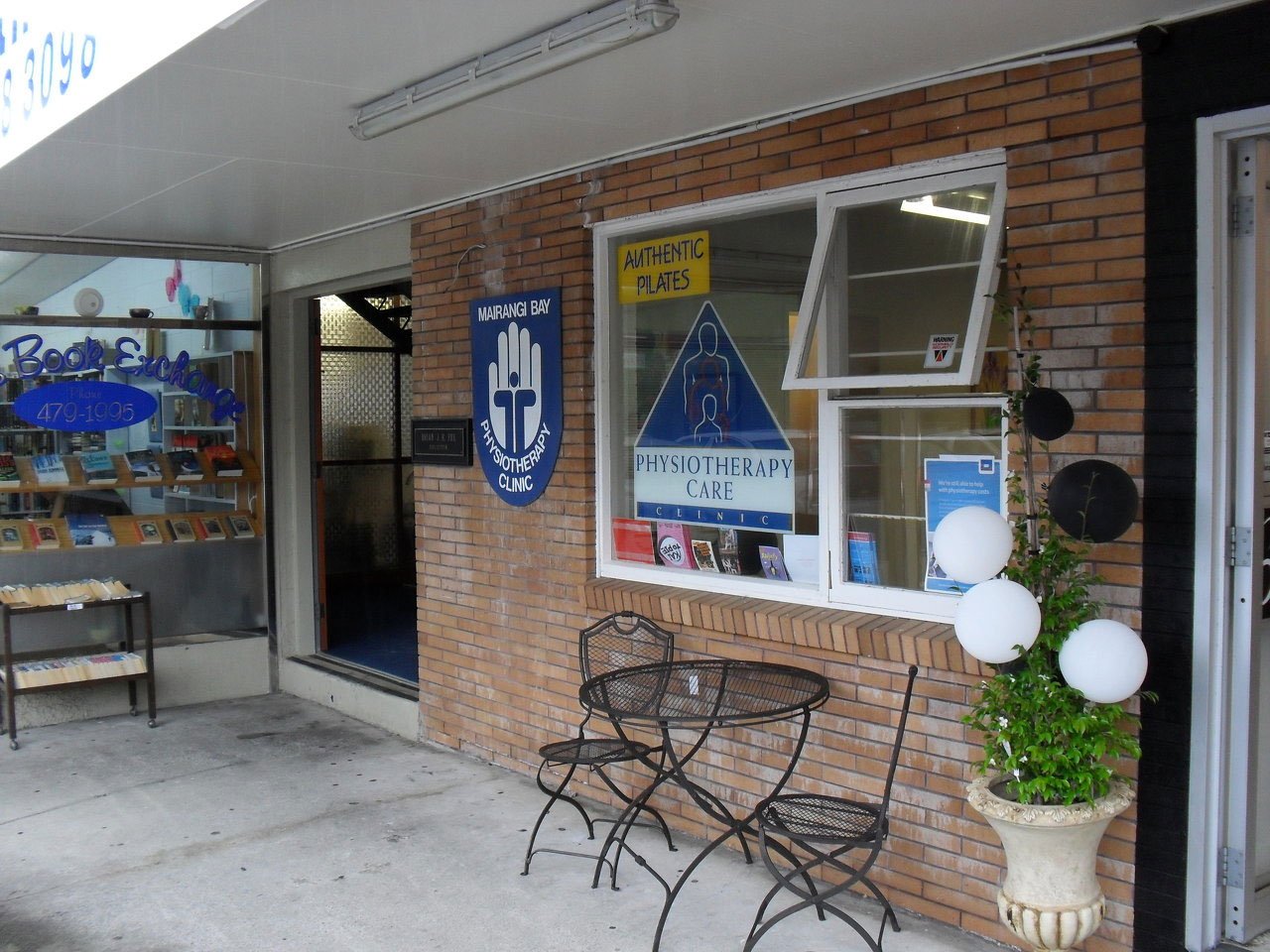 Exterior view of a physiotherapy clinic with a brick wall, window display of magazines, and outdoor seating including two black metal chairs and a round table. There is a large planter with a plant and white spherical lights, a sign reading 'Physiotherapy Care Clinic,' and another sign indicating 'Authentic Plates.' The windows display posters and books related to health and wellness.