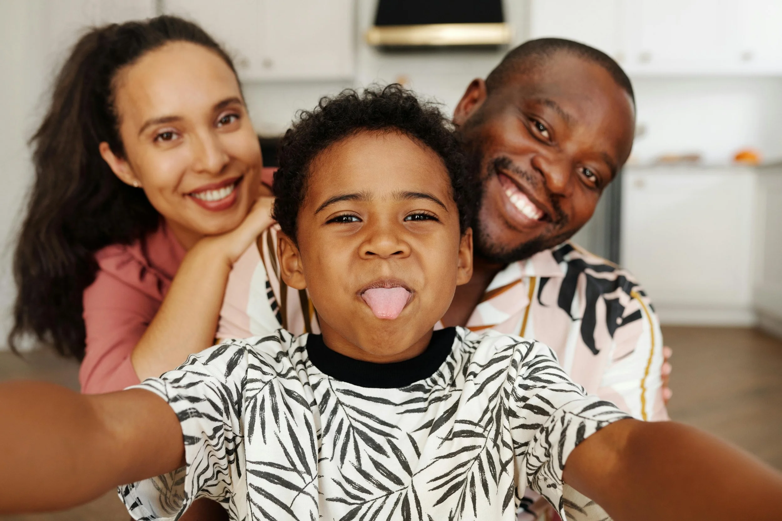 A family of three taking a selfie in their kitchen, with a woman and a man smiling and a young boy sticking out his tongue.