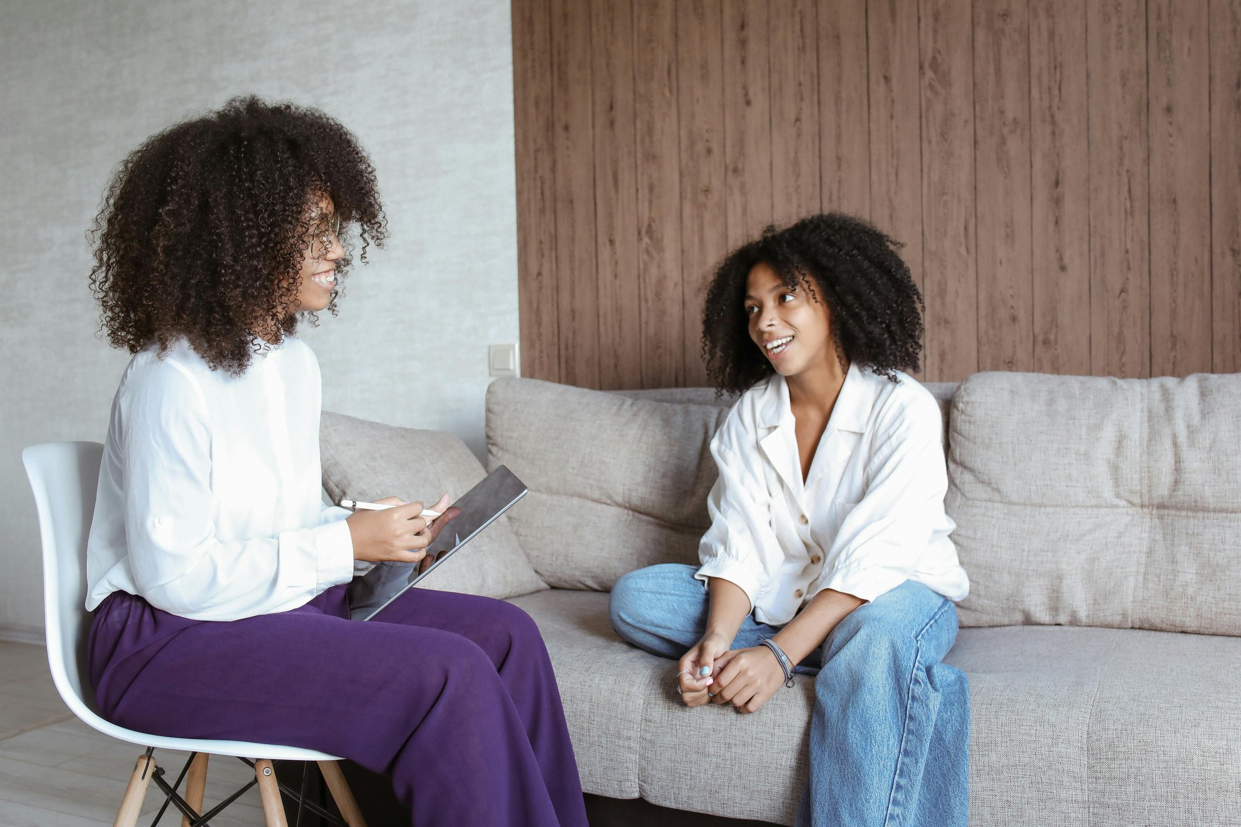 Two women with curly hair having a conversation in a living room. One woman is sitting on a chair with a notepad and pen, the other is sitting on a sofa, both smiling.