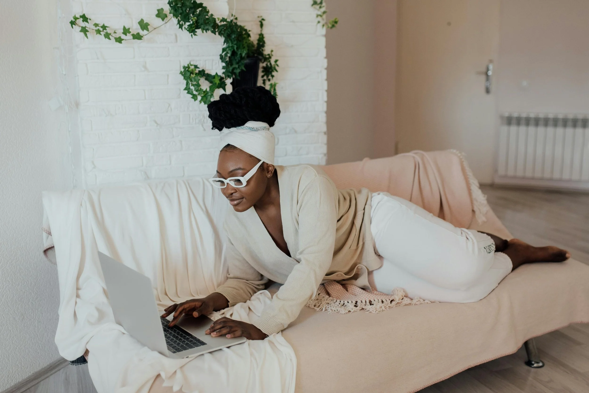 A woman with dark skin, wearing glasses and a white headwrap, is lying on her stomach on a couch, working on a laptop. She is dressed in a beige sweater and white pants, with a pink blanket draped over the couch; she looks focused on her work in a cozy living room with a white brick wall and green decorative plants.