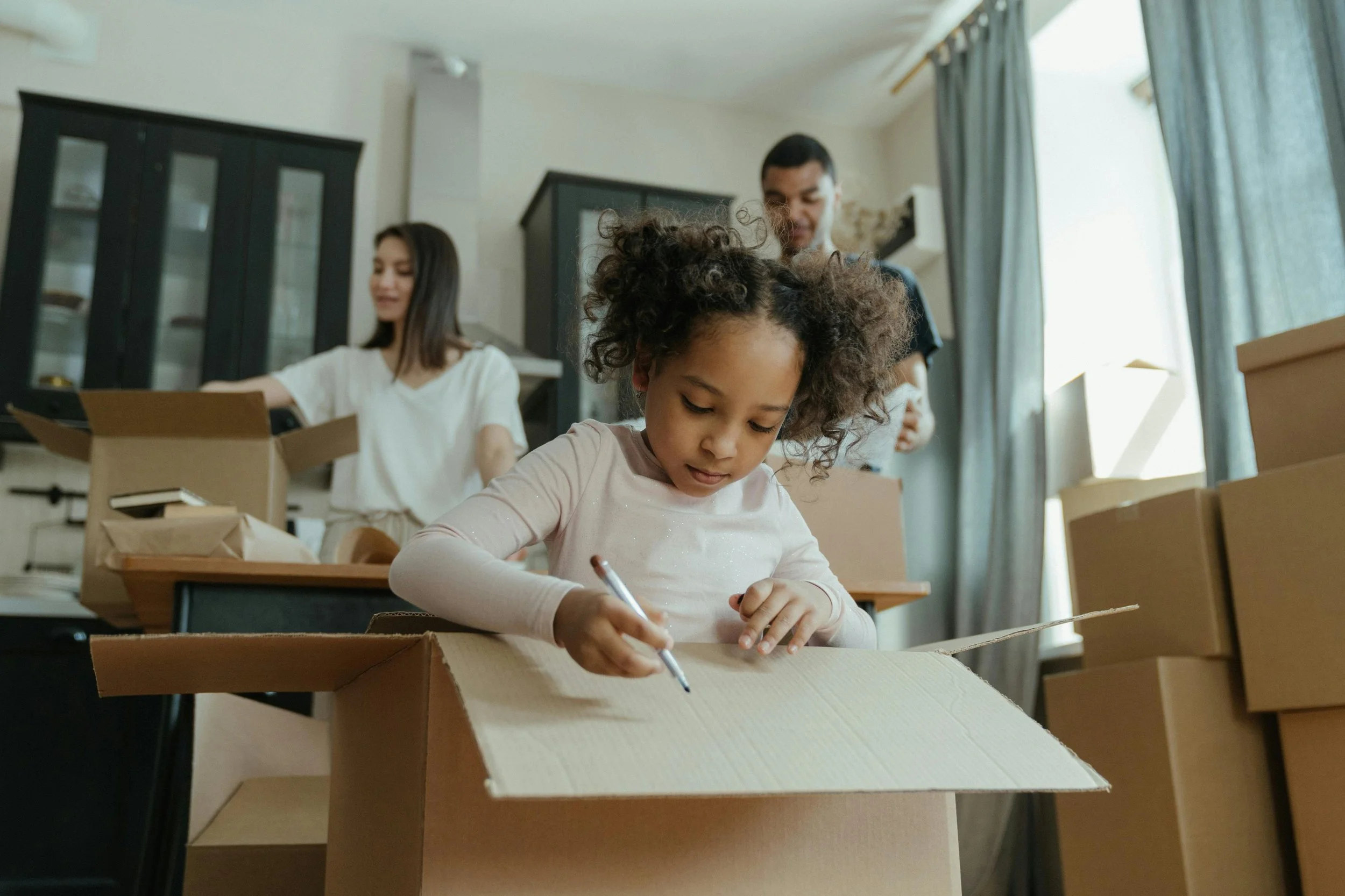 A young girl with curly hair writing on a cardboard box with a marker, in a room with unpacked boxes and a family in the background.