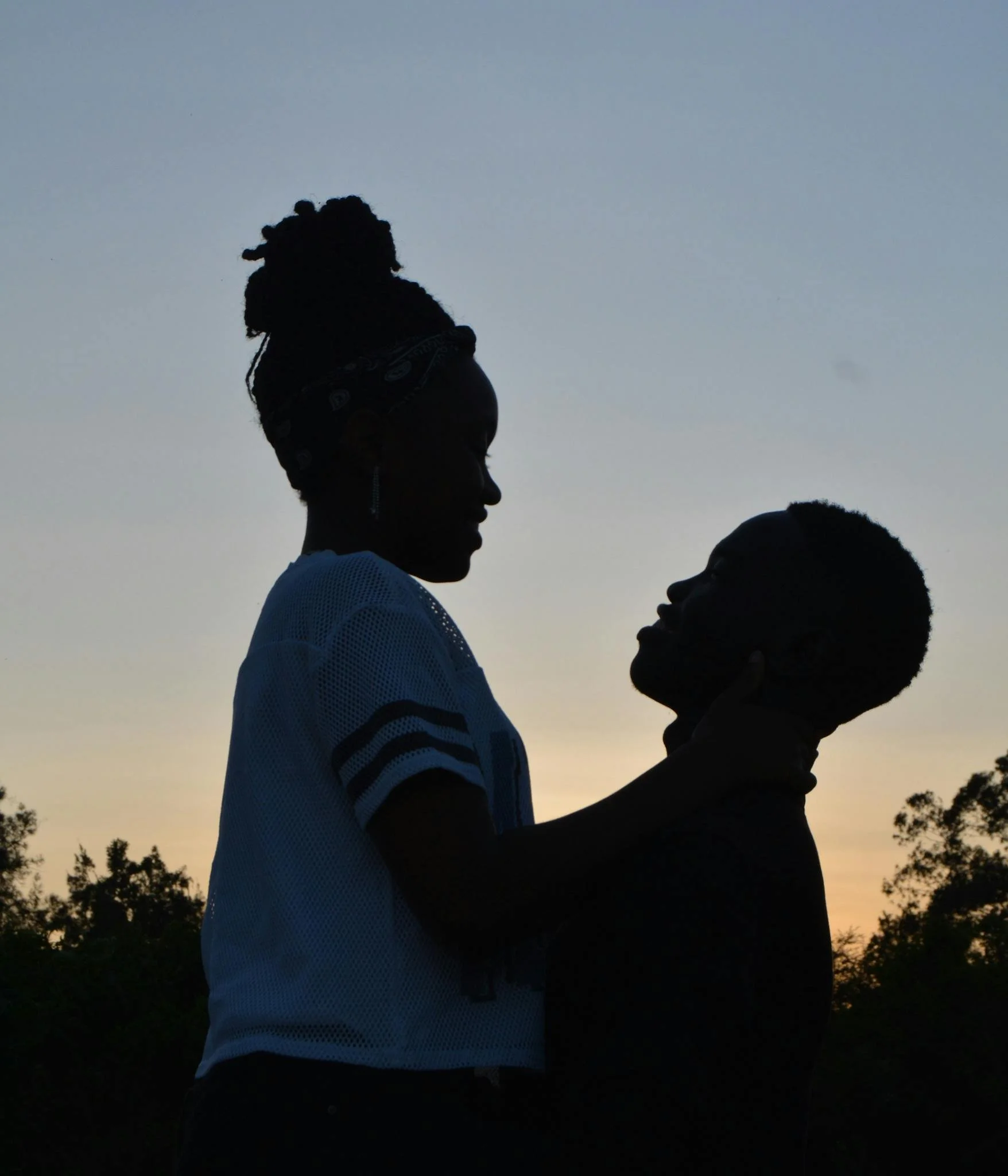 Silhouette of a woman and a man facing each other at sunset, with a clear sky and trees in the background.
