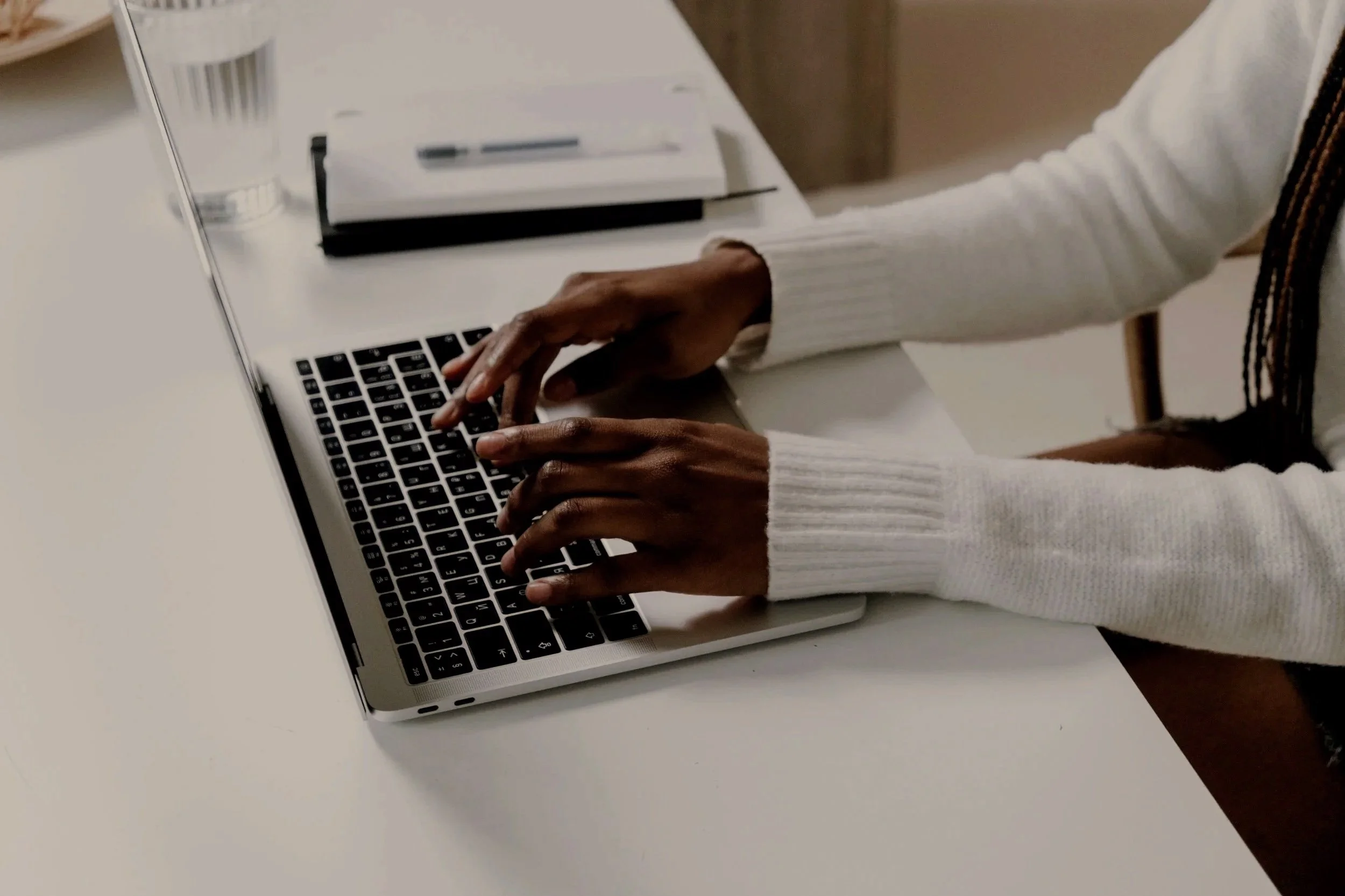 Person typing on a silver laptop keyboard at a white desk with a glass of water, a notepad, and a pen.