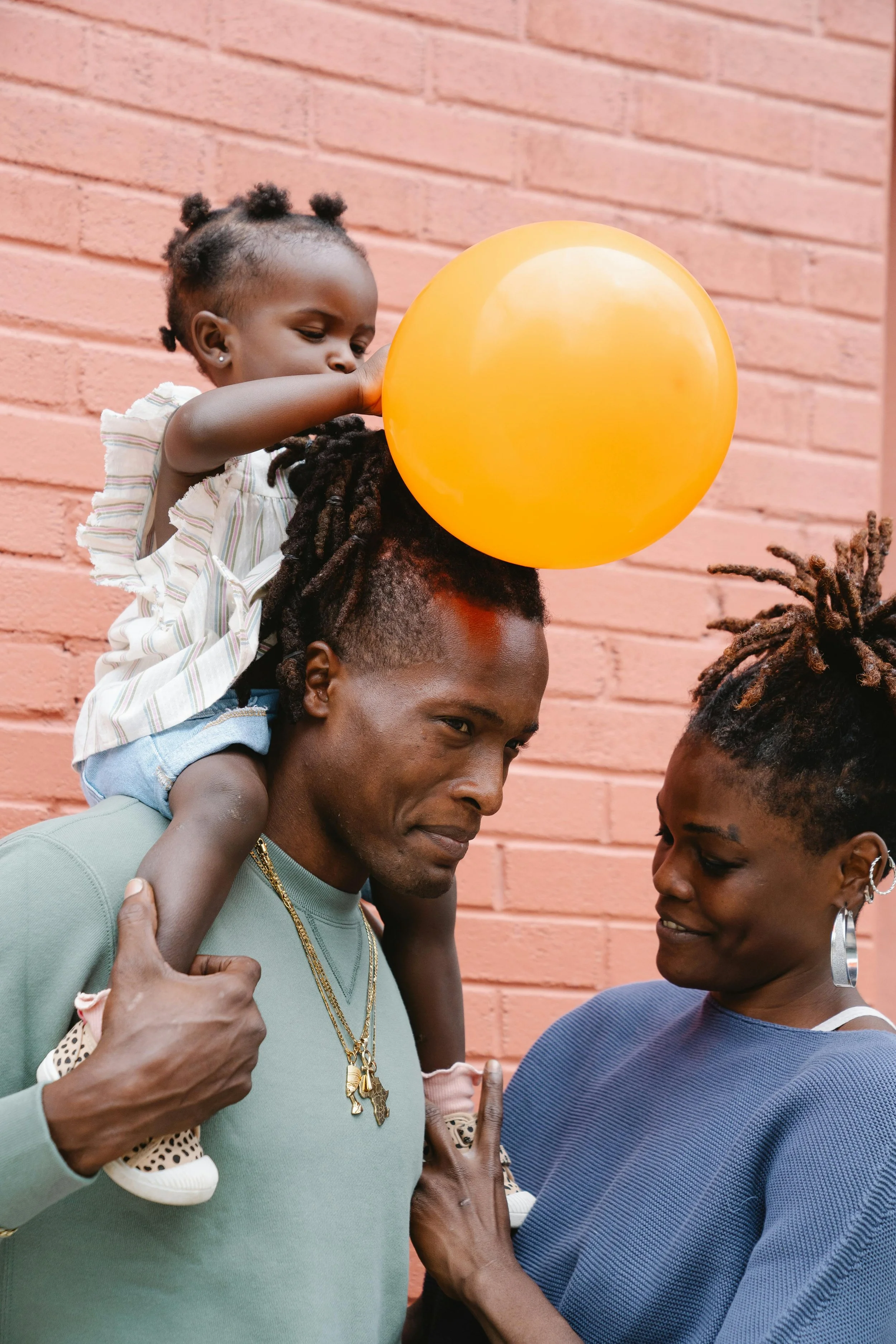 A family of three smiling together outdoors near a brick wall. The father has reddish dreadlocks and wears a green shirt with layered necklaces. The mother has short textured hair with hoop earrings and wears a blue top. The young girl, with puffy hair in small buns, is sitting on the father's shoulders holding an orange balloon.