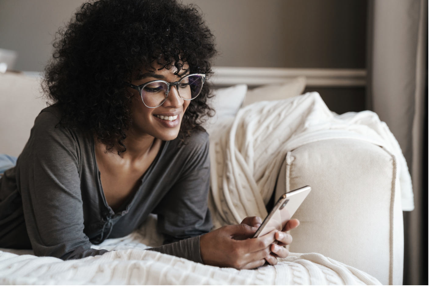 A woman with curly hair and glasses smiling while looking at her smartphone in bed.