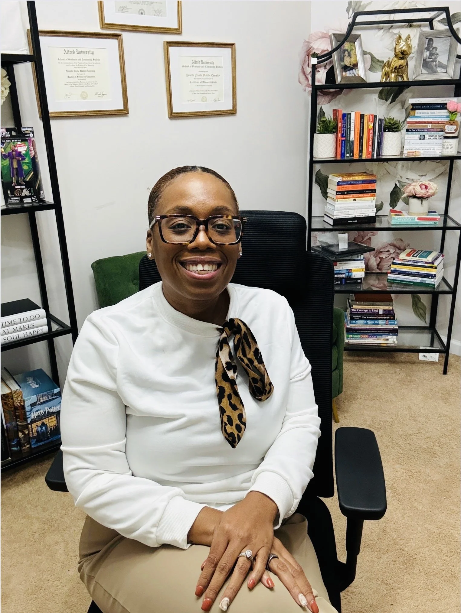 A smiling woman with glasses and a white top with a leopard print scarf, seated in an office with shelves of books and framed certificates.