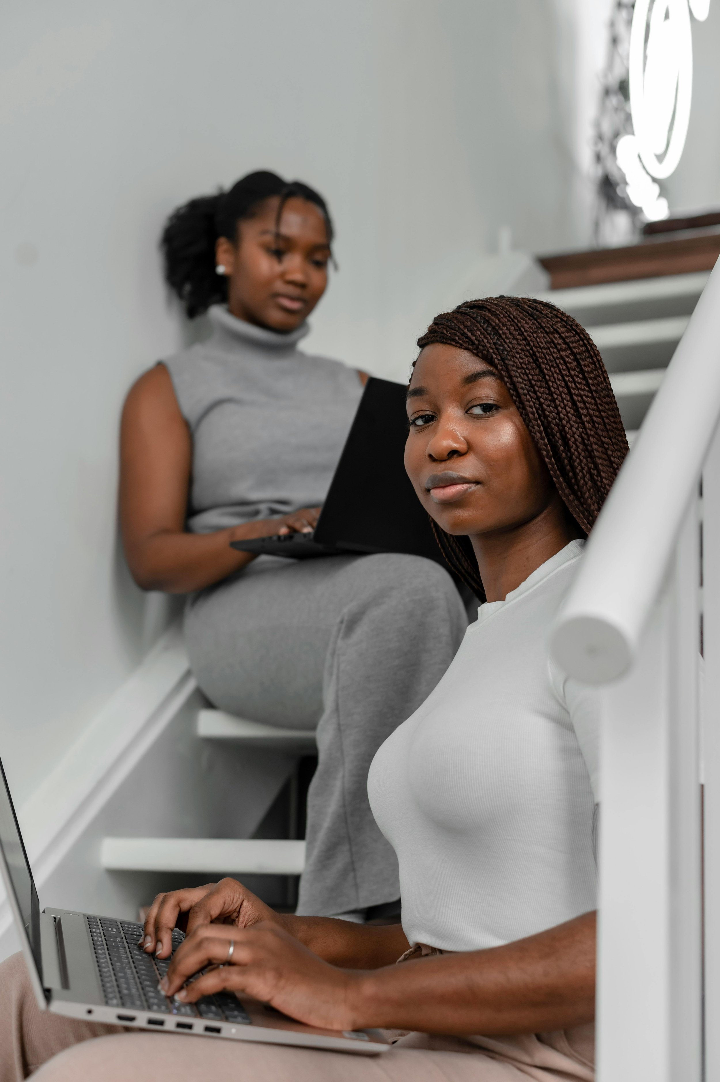 Two women working on laptops on a staircase, one sitting on the step below and the other sitting on the staircase, both looking at the camera.