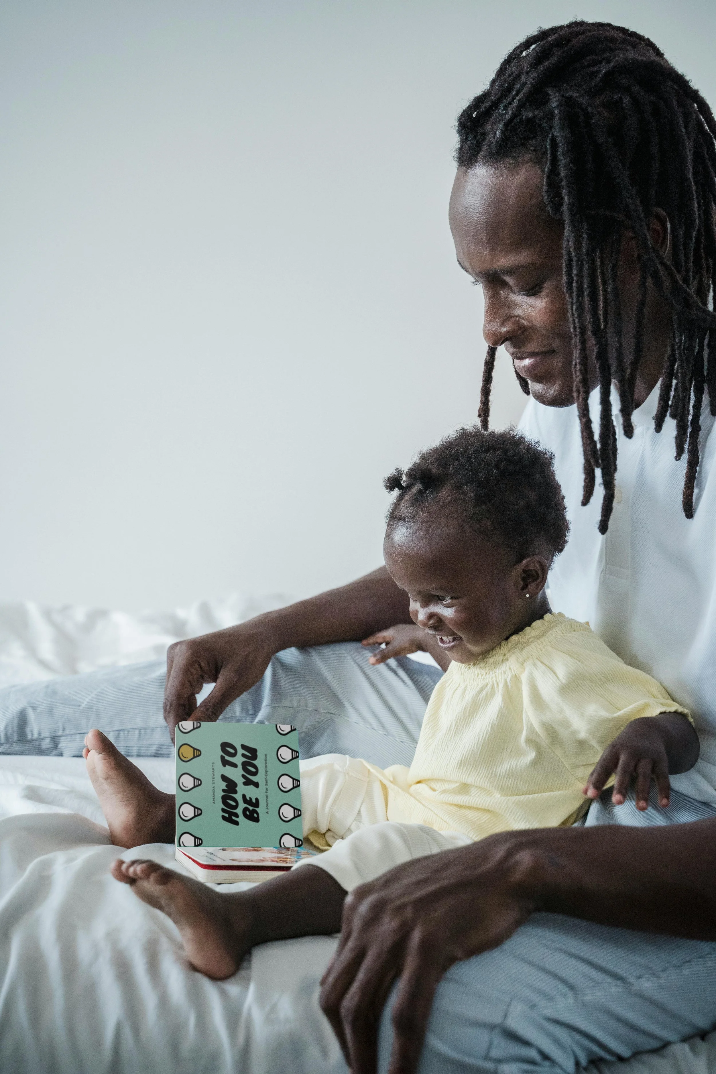 A man and young girl sitting on a bed looking at a book with the words 'How to Be You' on the cover, smiling together.