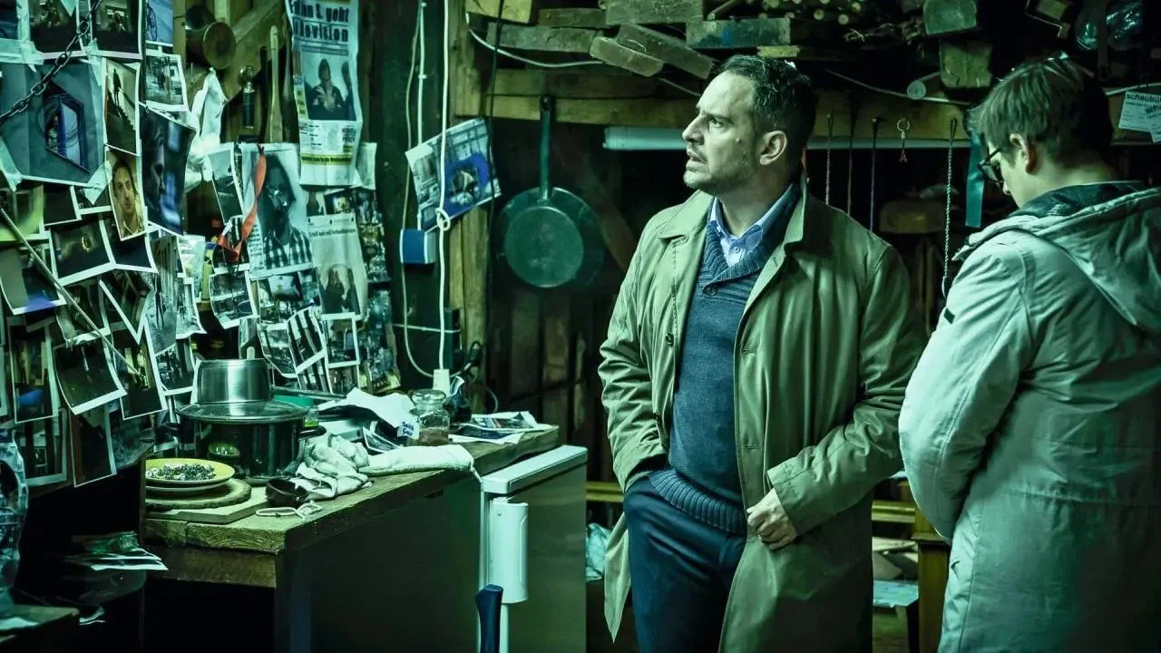 Paul is looking at the photos and articles on the wall in disbelief. Ingolf is also in the photo looking at his phone.