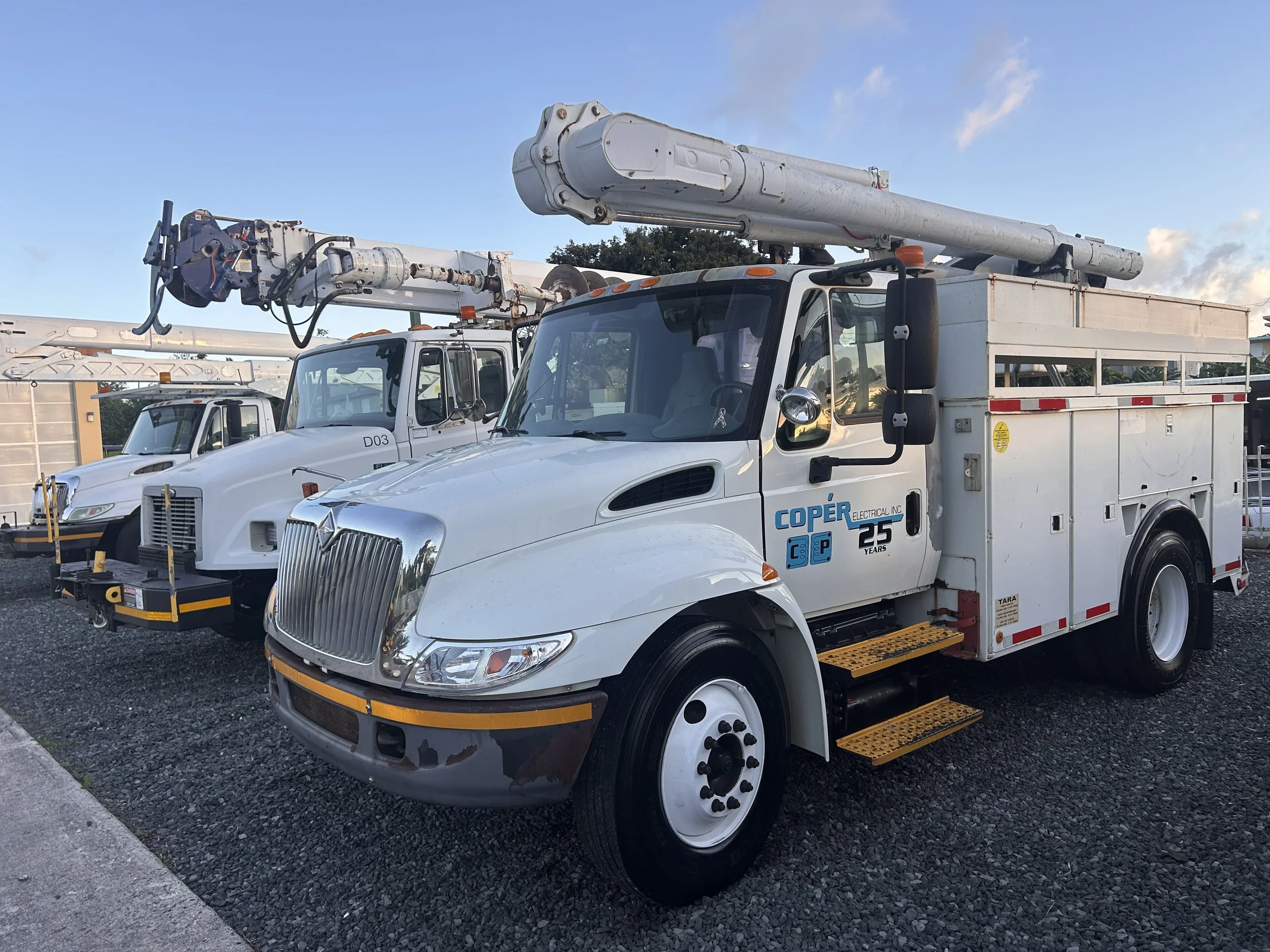 Two utility bucket trucks parked side by side on a gravel lot, with the sky partly cloudy in the background. The truck in the foreground has a logo that reads 'Coper Electrical Inc' on the door.