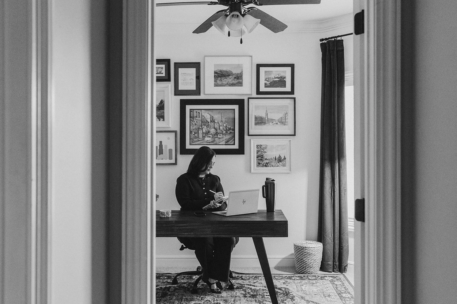 A woman sitting at a desk, looking at her phone, with a laptop open in front of her. The room has a gallery wall with framed pictures and paintings, a window with curtains, a basket on the floor, and a ceiling fan.