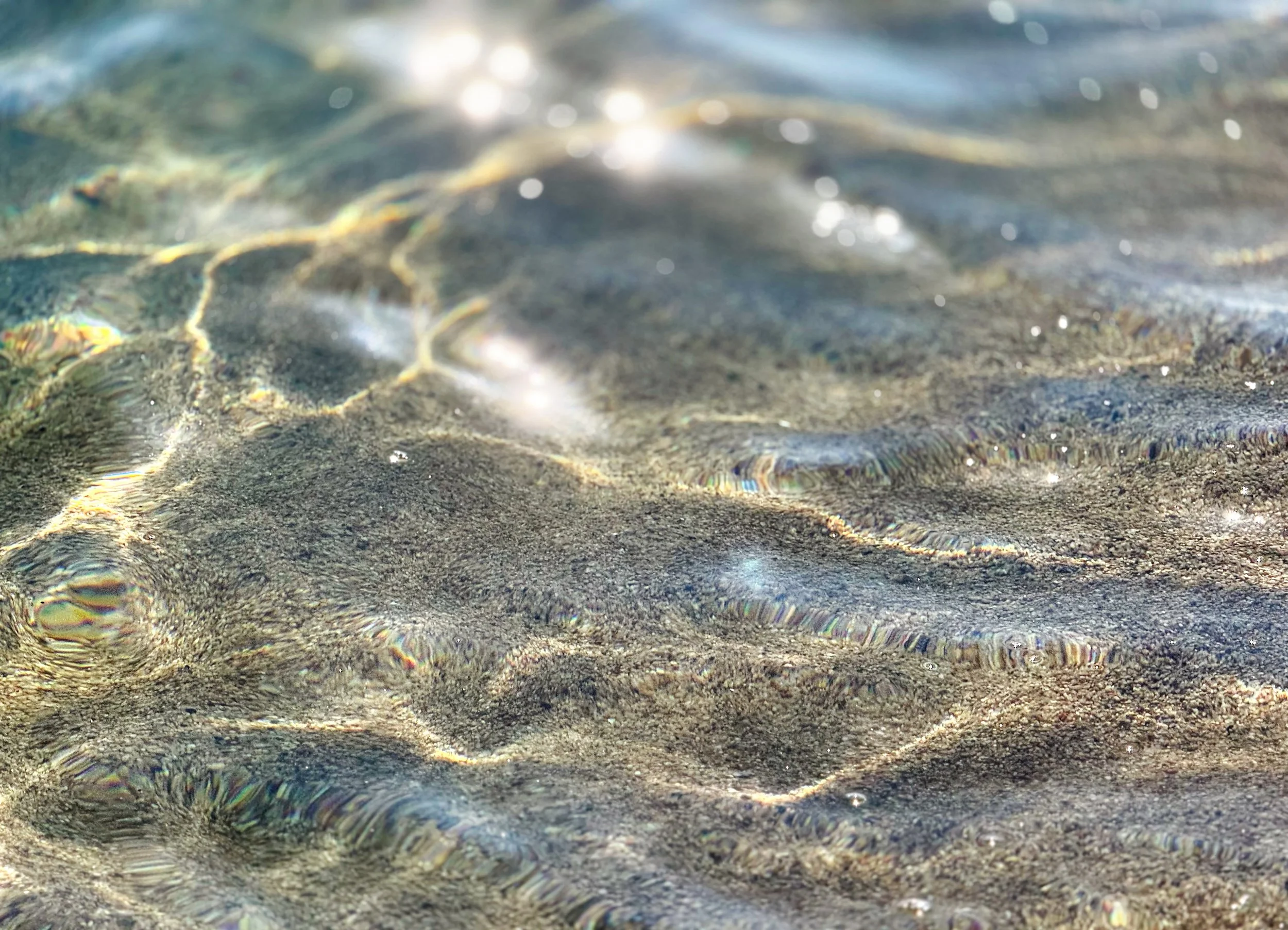 Close-up of clear water and sand at the beach, with sunlight reflecting on the surface.  Signifying clarity and depth.