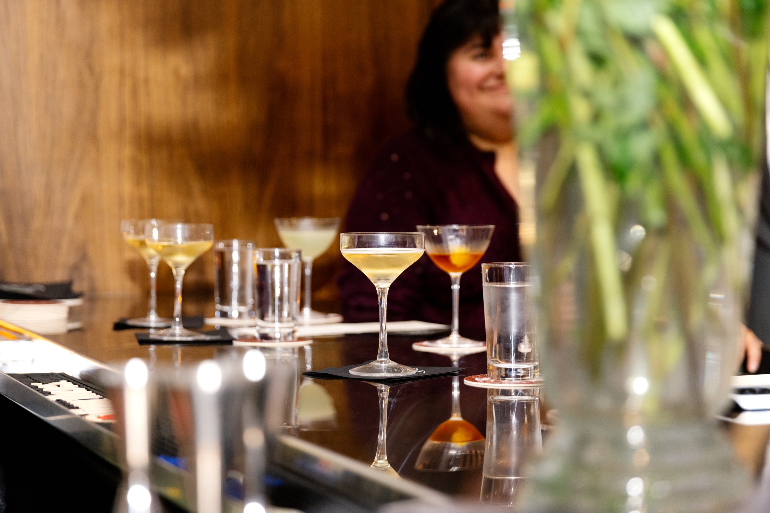 A woman smiling in the background at a bar or restaurant table with various cocktail glasses and a tall glass with water in the foreground.