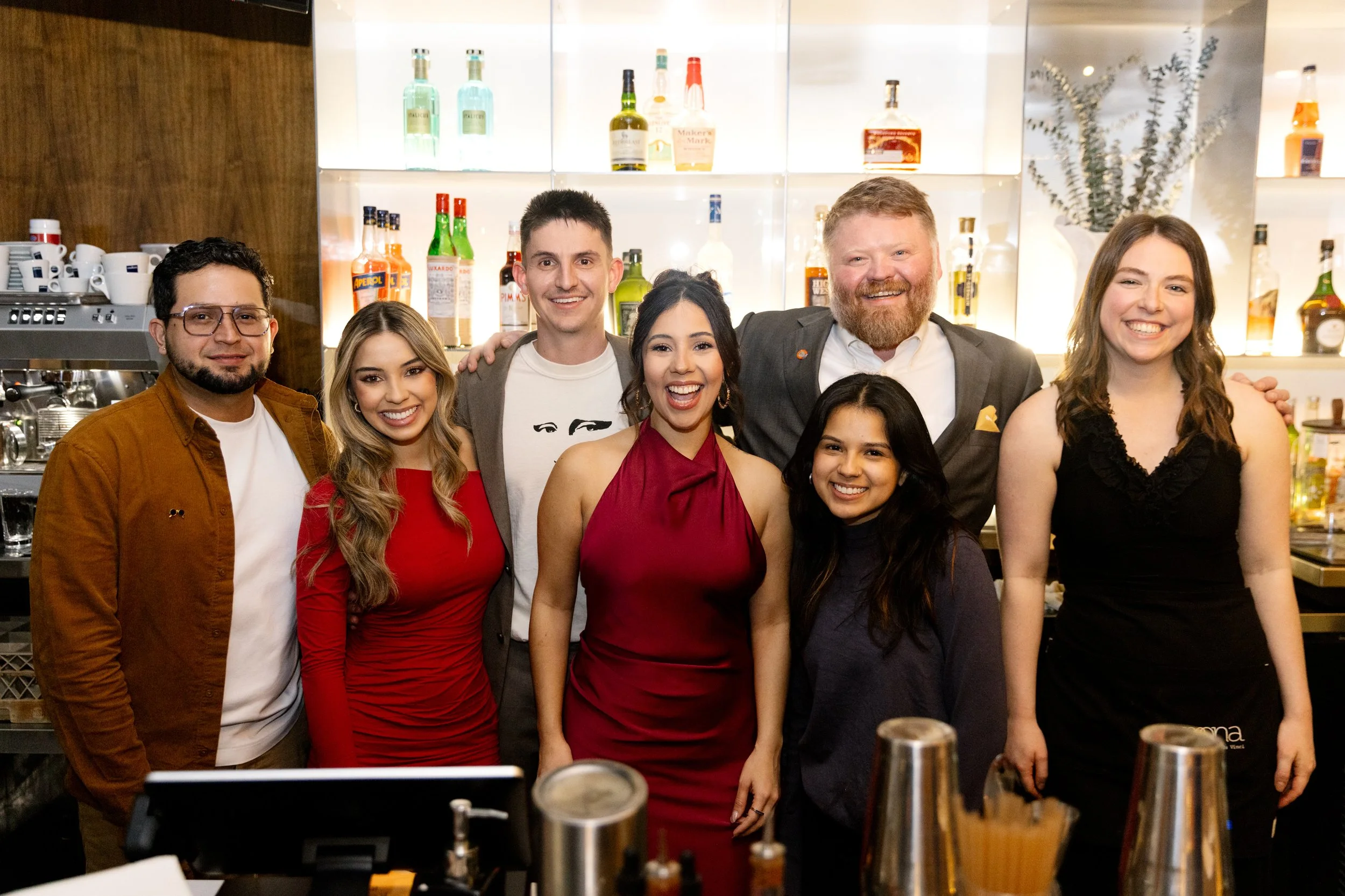 Group of seven diverse people smiling in a bar or restaurant, with bottles and bar equipment in the background, celebrating together.