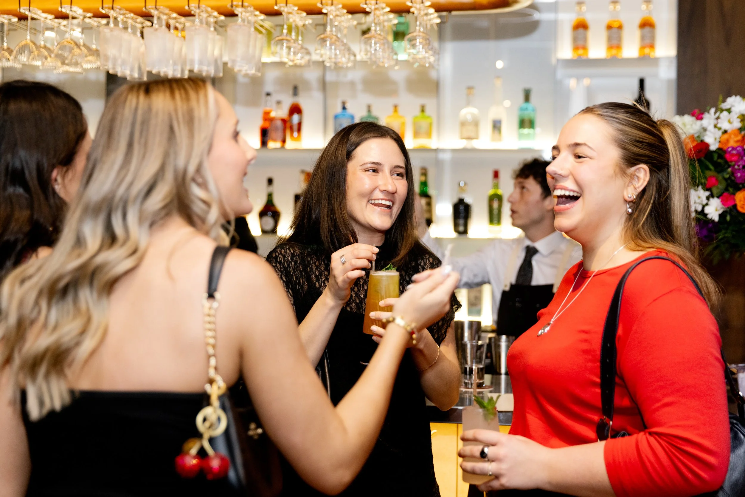 Group of women laughing and enjoying drinks at a bar with colorful bottles and flowers in the background.