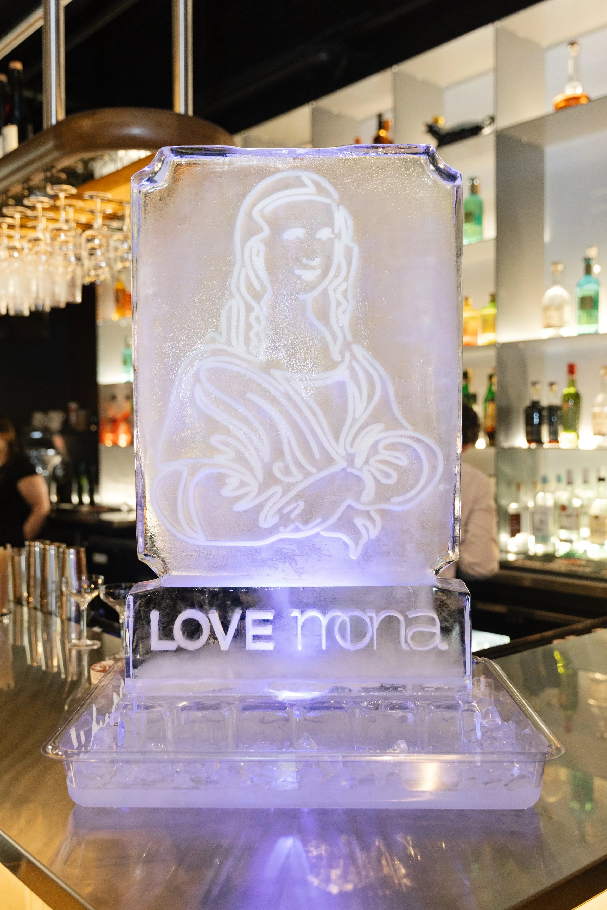Ice sculpture featuring Mona Lisa's face and the words 'LOVE Mona' beneath it, illuminated with purple light, in a bar setting.