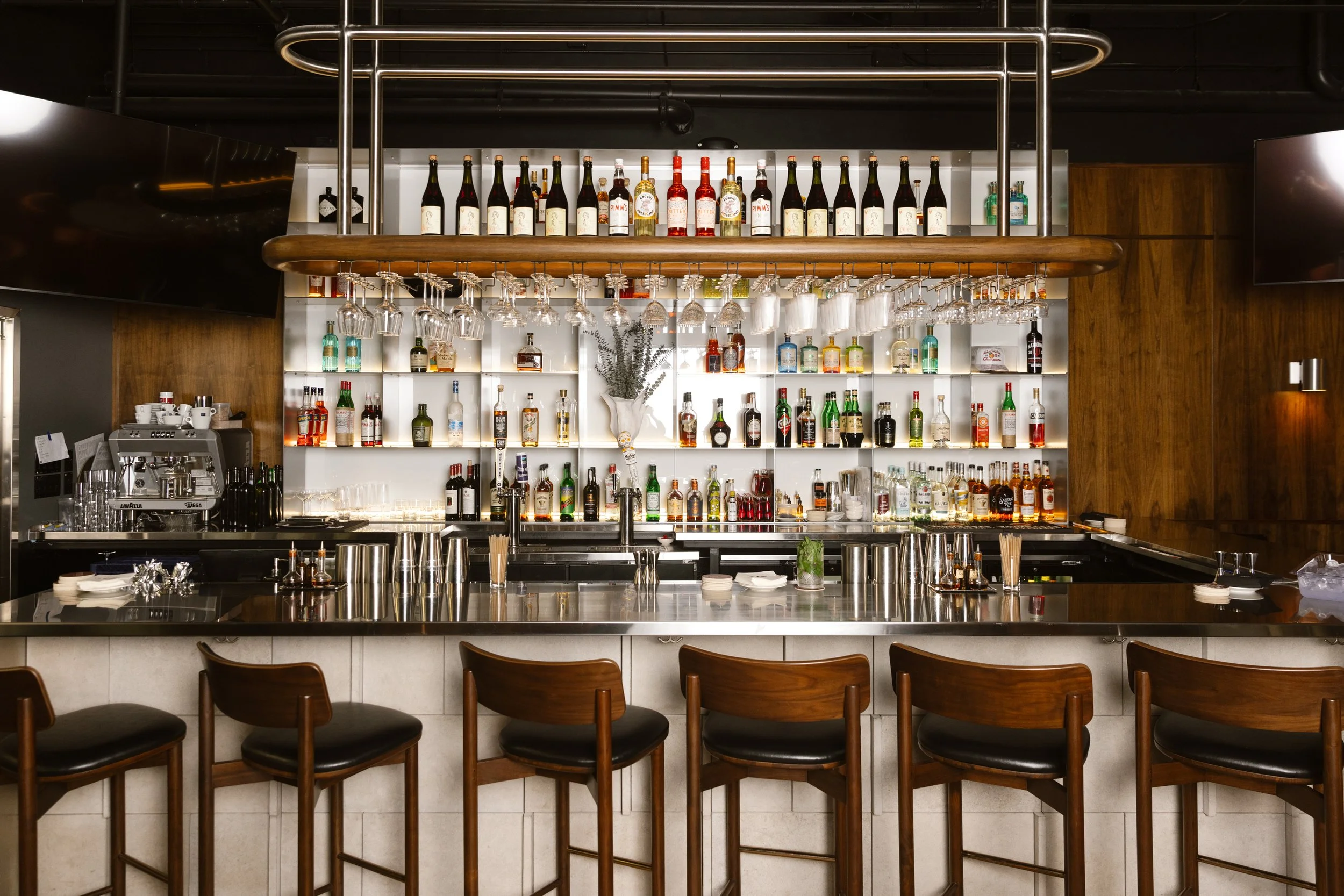 A bar with a variety of bottles of alcohol, hanging wine glasses, a coffee machine, and bar stools.