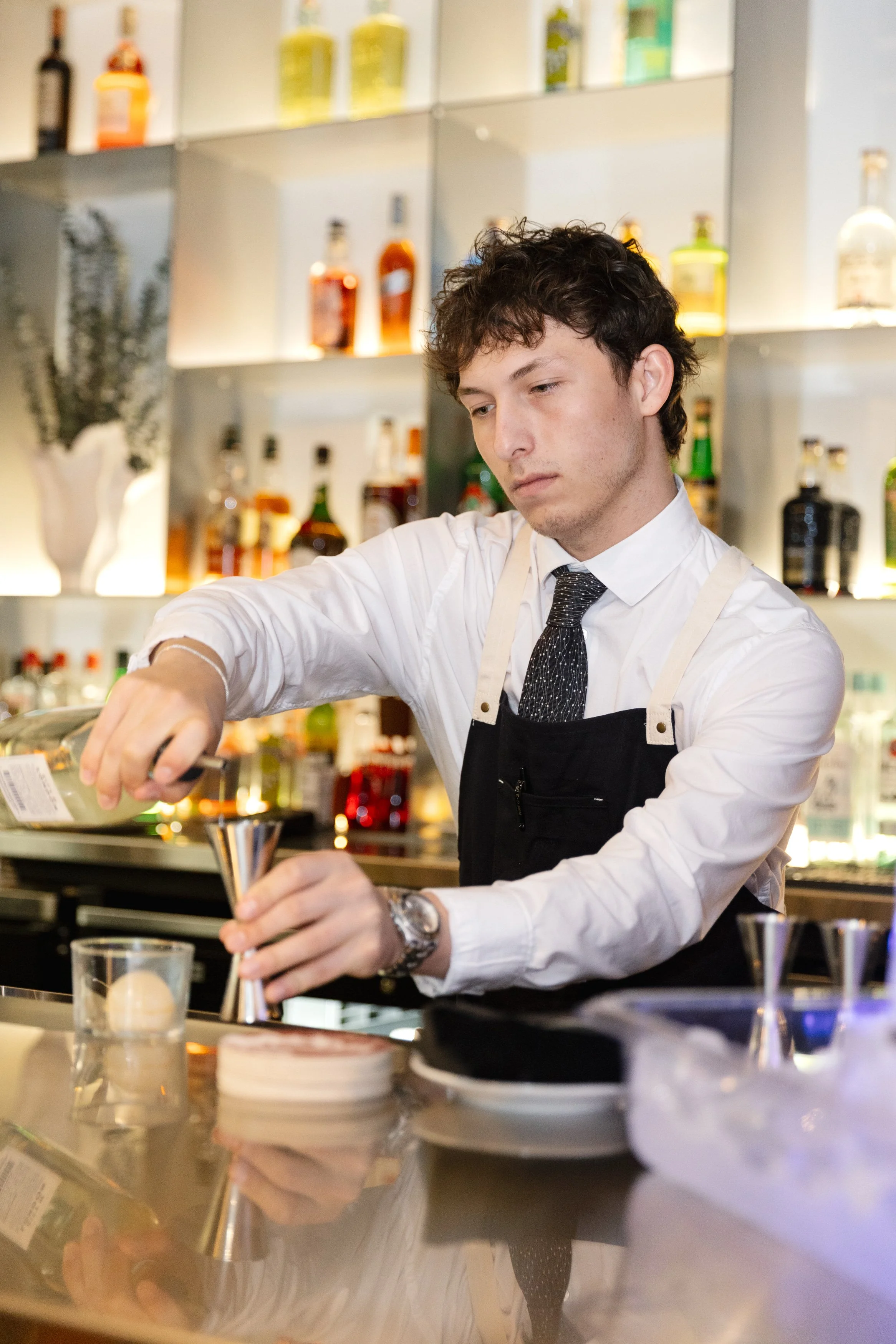 A bartender in a white shirt, black apron, and tie preparing a cocktail at a bar with bottles of alcohol on shelves in the background.