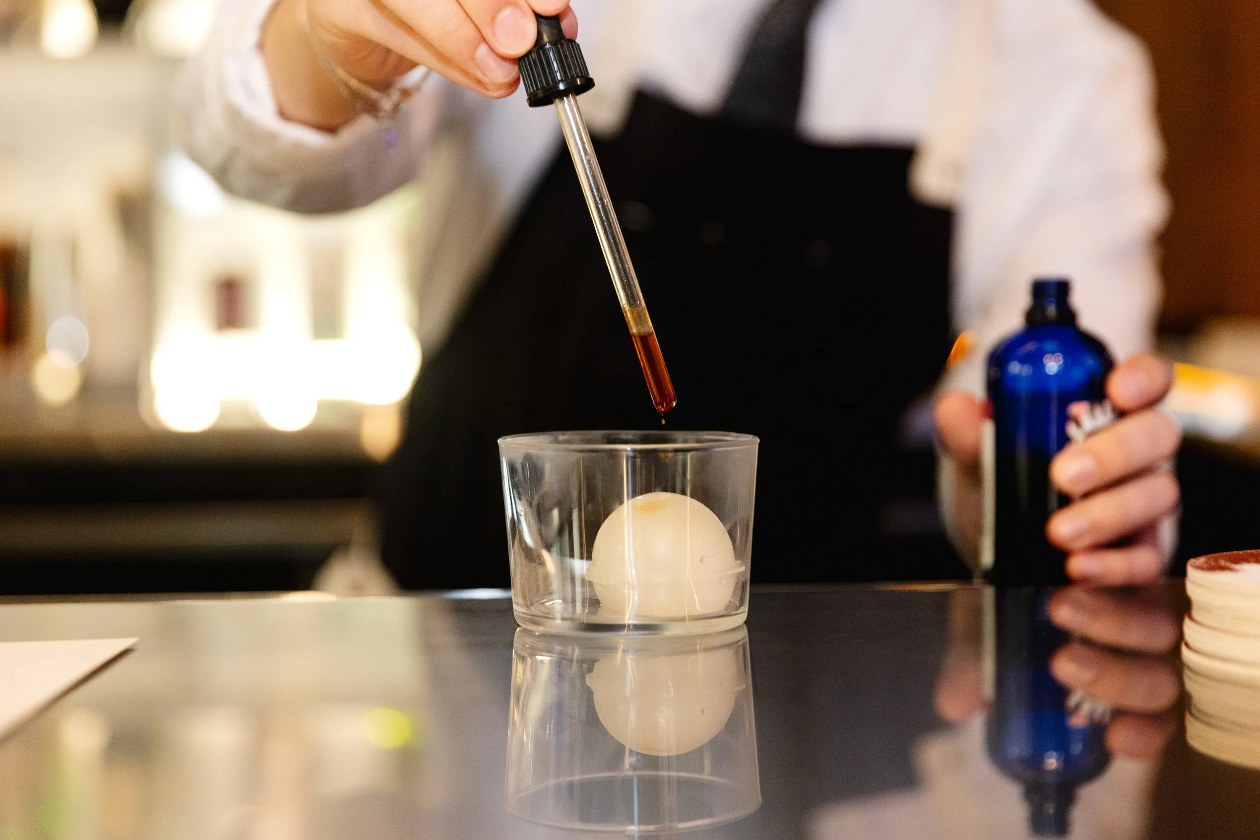Person adding a liquid using a dropper to a clear glass with a white sphere inside, on a reflective surface.