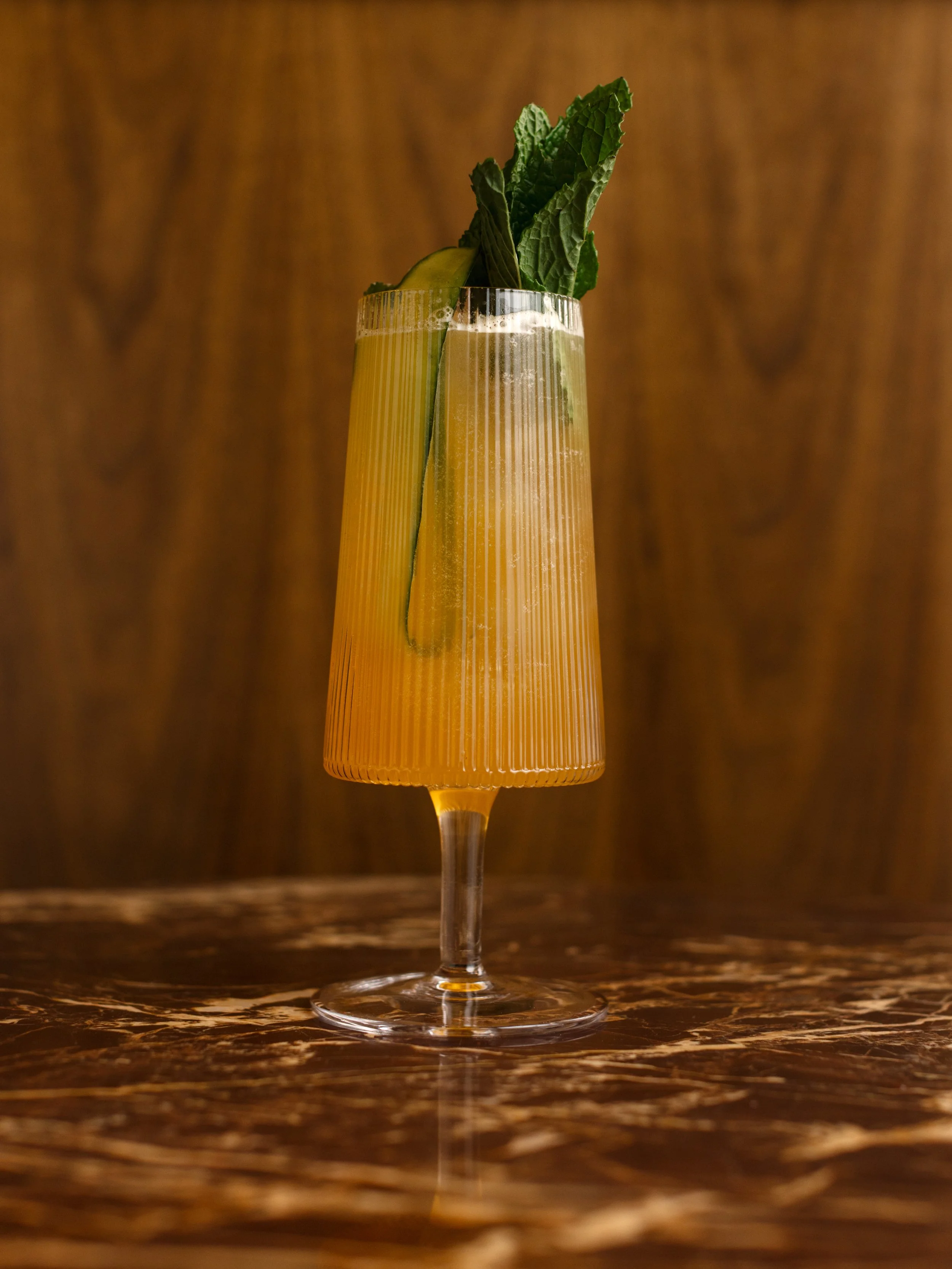 A tall glass of yellow-orange cocktail garnished with mint leaves, a cucumber slice, and a lemon wedge, set on a dark marble surface with a wooden background.