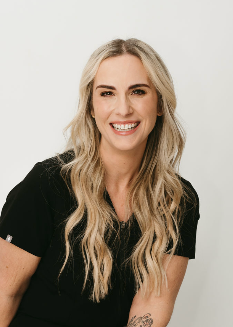 A smiling woman with long blonde wavy hair wearing a black top against a plain white background.