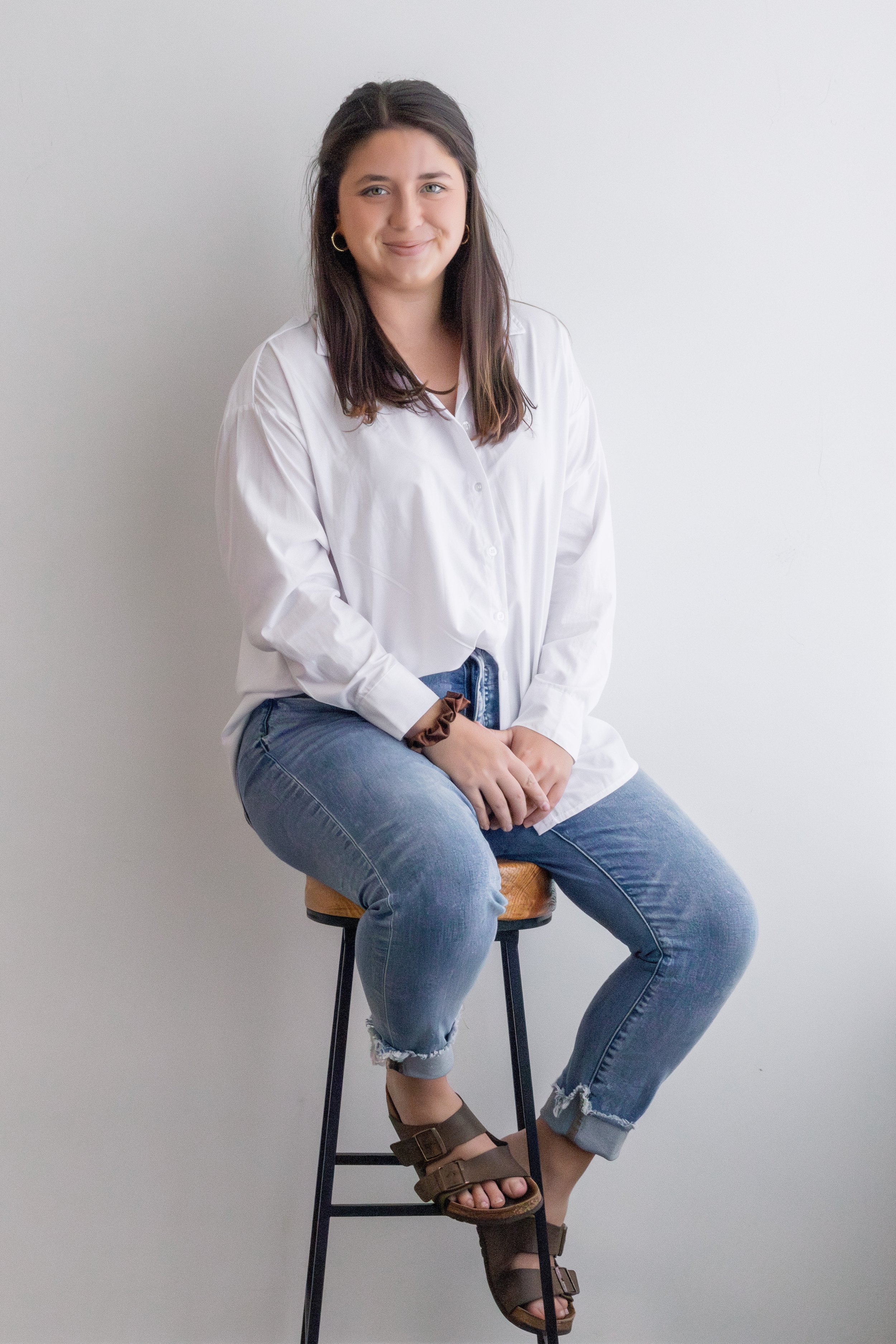 A young woman sitting on a wooden stool against a plain white wall, wearing a white button-up shirt, blue ripped jeans, and brown sandals, smiling at the camera.