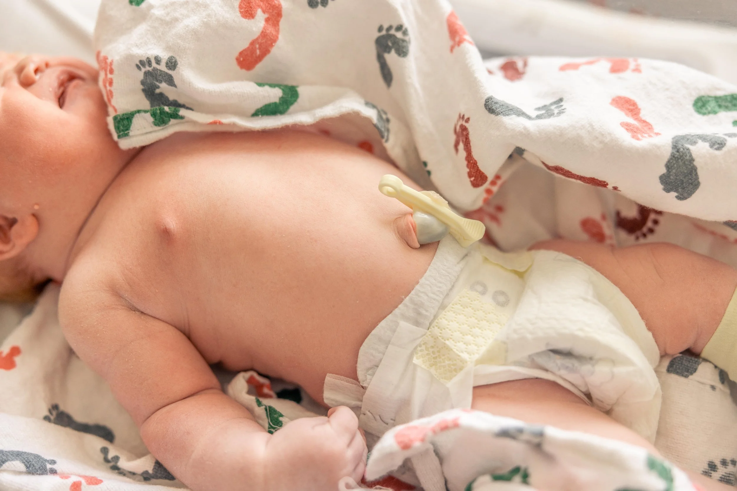 A newborn baby lying on a hospital bed, wrapped in a blanket with multicolored footprints, wearing a diaper and connected to a medical cord.