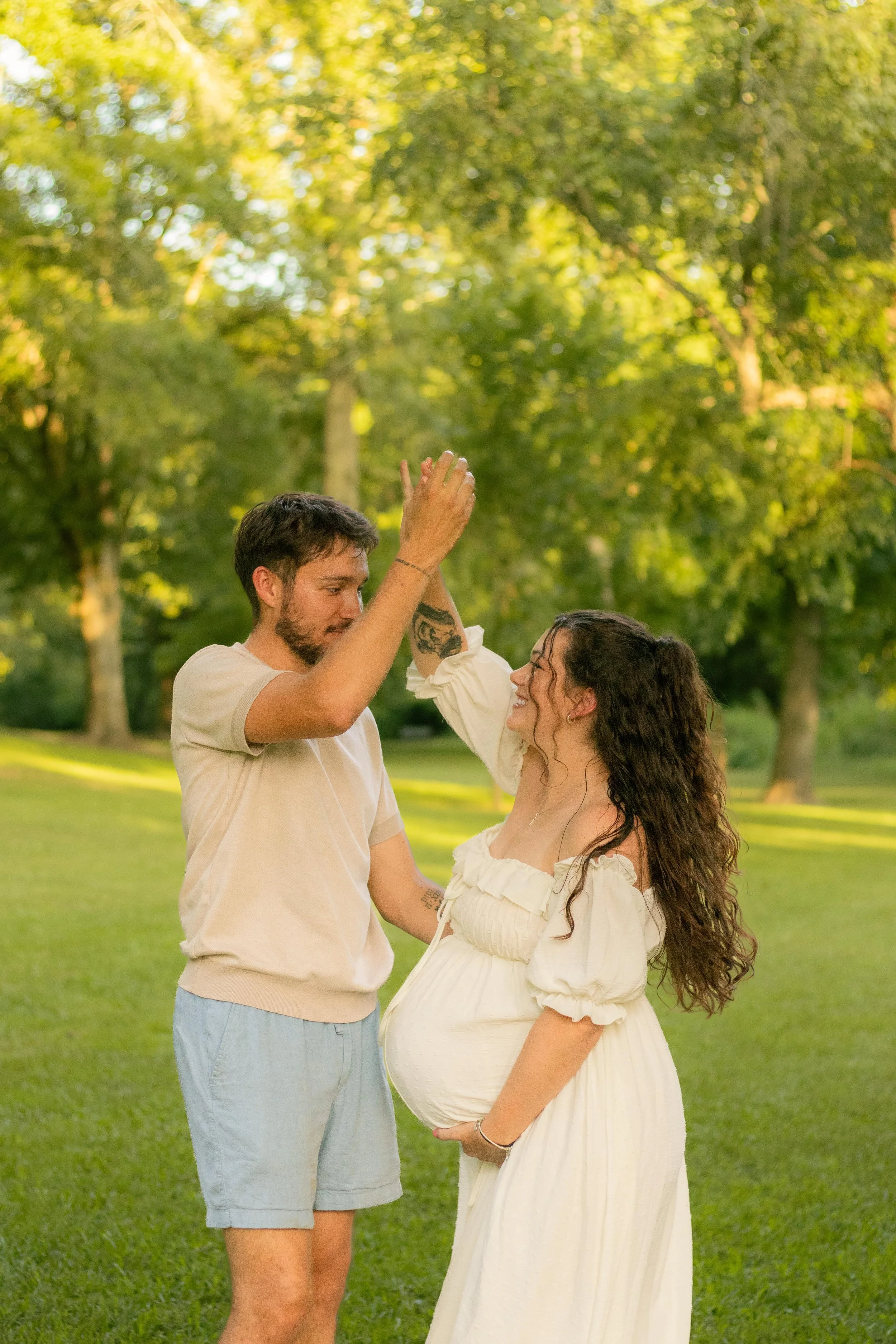 A pregnant woman in a white dress and curly hair smiling as a man in casual clothing gives her a high five outdoors in a park with green trees.