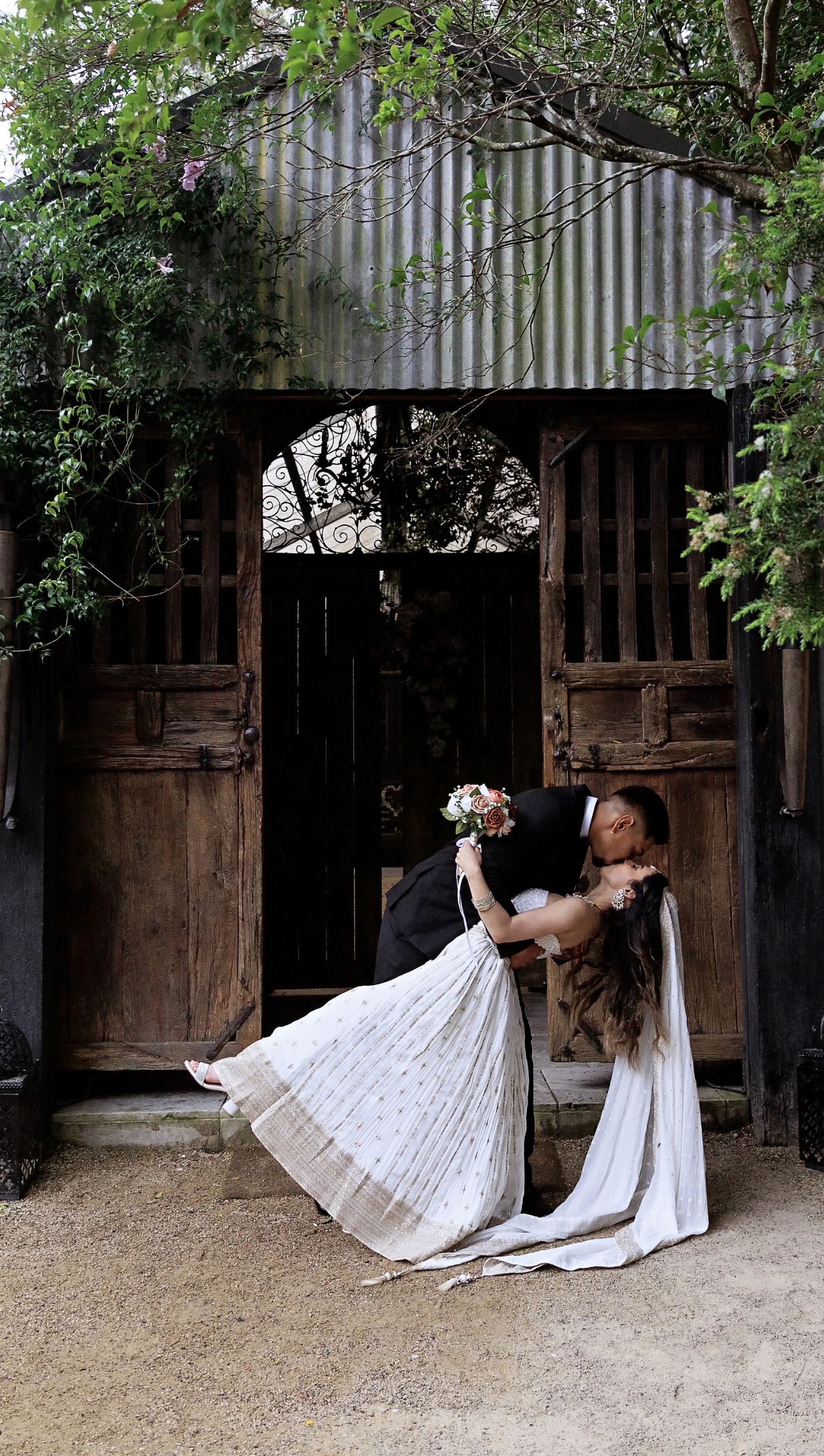 A couple dressed in wedding attire sharing a romantic kiss outside a wooden gate surrounded by greenery.