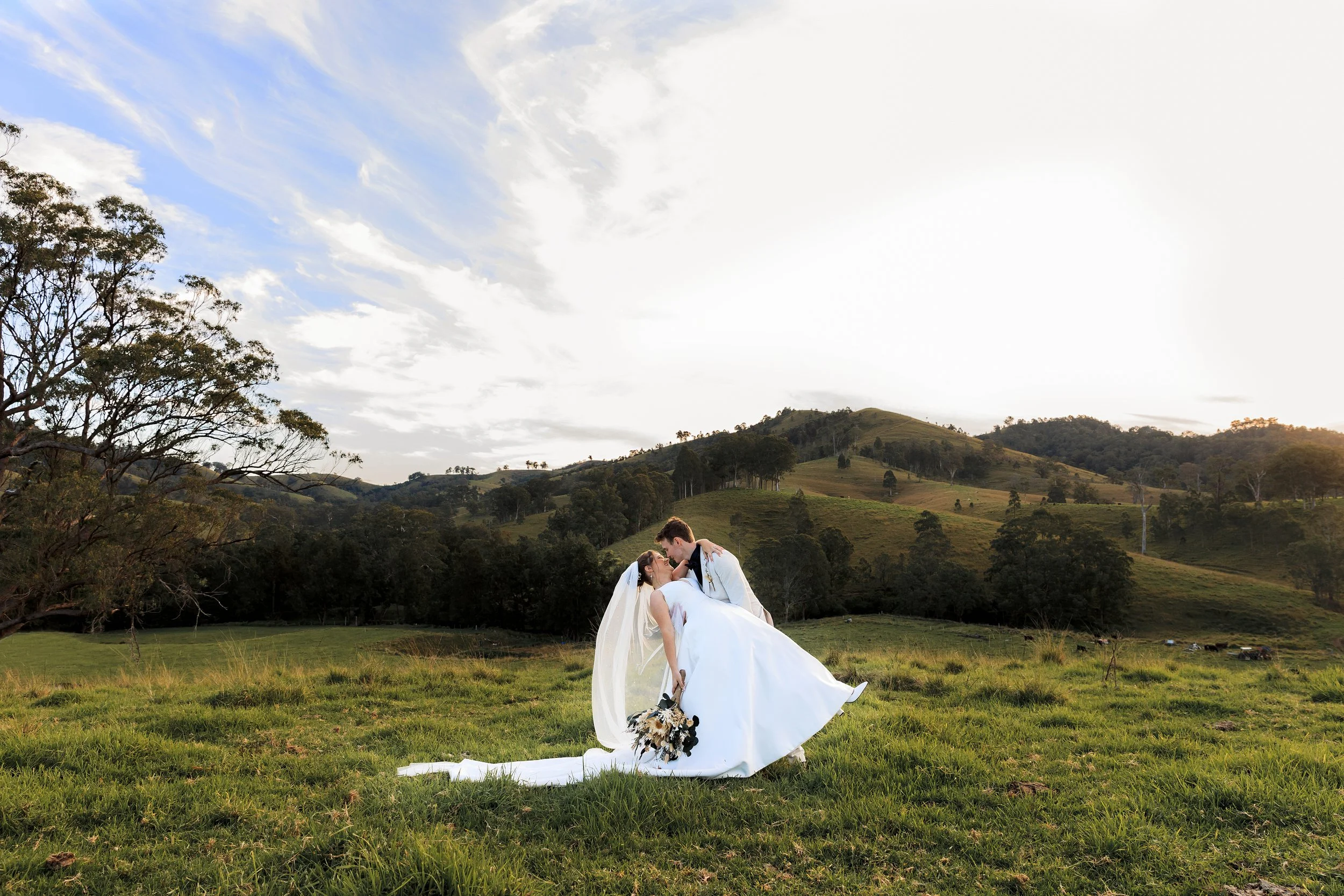 A couple in wedding attire the groom dips the bride holding a bouquet of flowers in a grassy field with rolling hills, trees, and a partly cloudy sky during sunset.