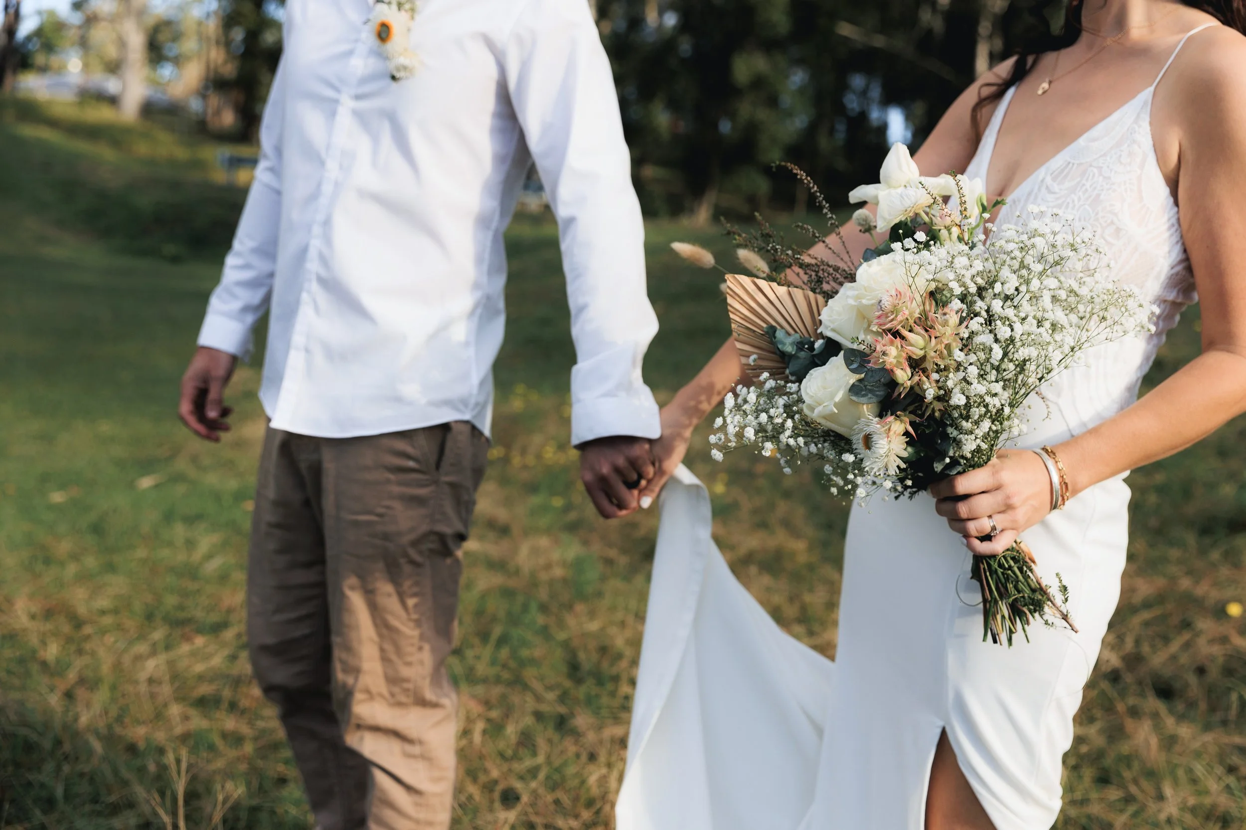 A bride holding a bouquet of white flowers and greenery, standing outdoors with a man, both holding hands. The woman's dress is white with thin straps, and the man's attire includes a white shirt and tan pants. The background features blurred trees a
