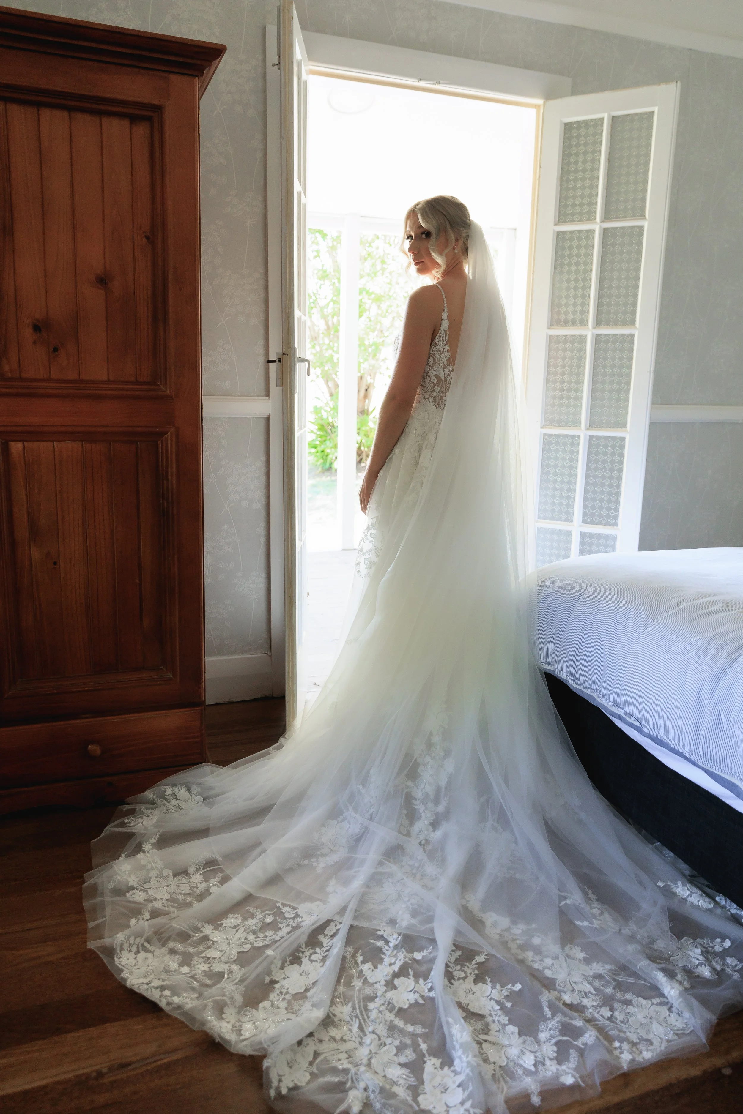 A bride in a wedding dress with a long, embroidered train, standing by an open door that leads outside to greenery. The dress features lace details on the back and she has a veil. The room has wooden flooring and a wooden wardrobe on the left.