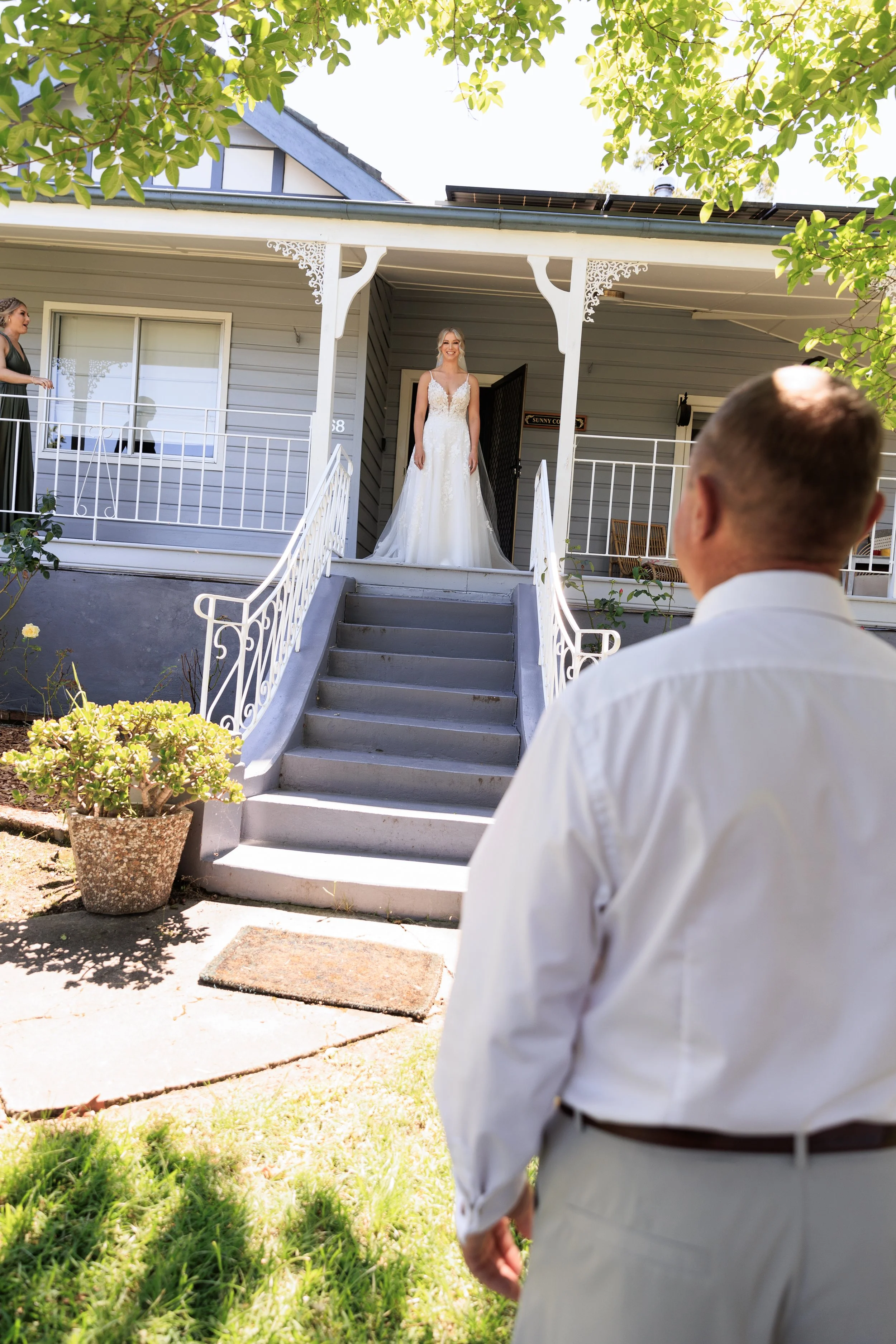 A man in a white shirt and beige pants is walking up the steps to a house. There is a woman in a black dress on the porch. A bride in a white wedding dress is standing at the door, smiling. The house is light blue with white trim, and there are trees
