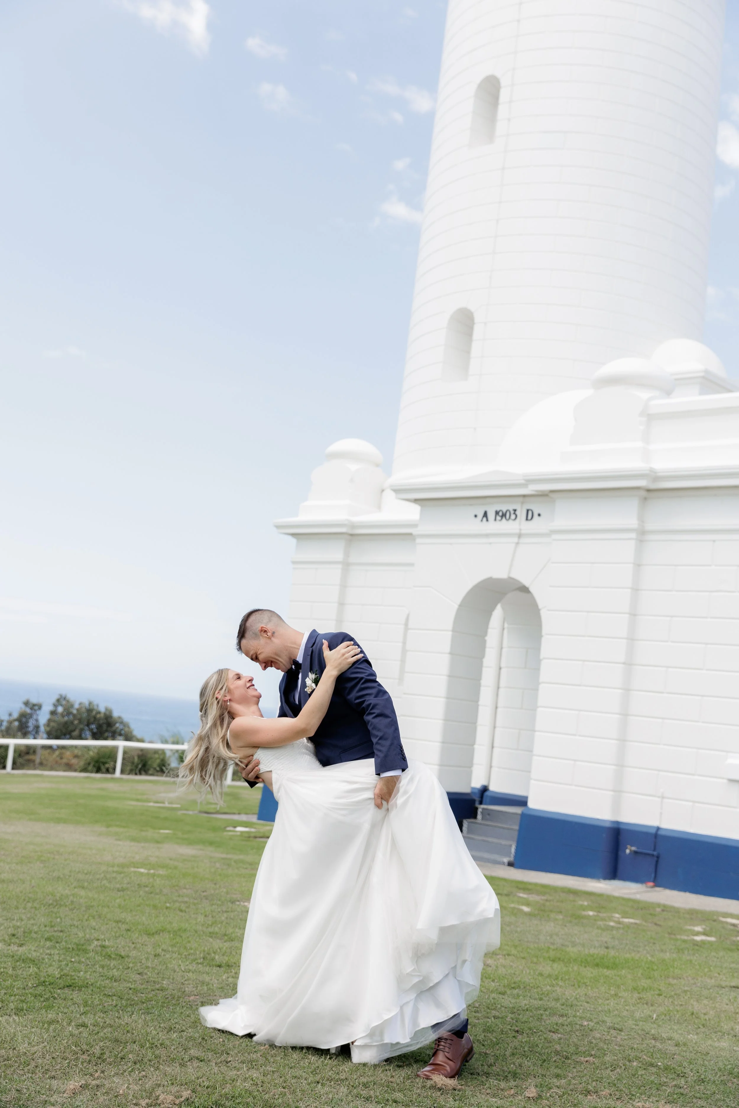 A newlywed couple, with the groom in a navy suit and brown boots and the bride in a white wedding dress, are enjoying a joyful moment outdoors in front of a white lighthouse during daytime. The groom is lifting the bride who is smiling and holding onto him, with a scenic background of cloudy sky, distant ocean, and green grass.
