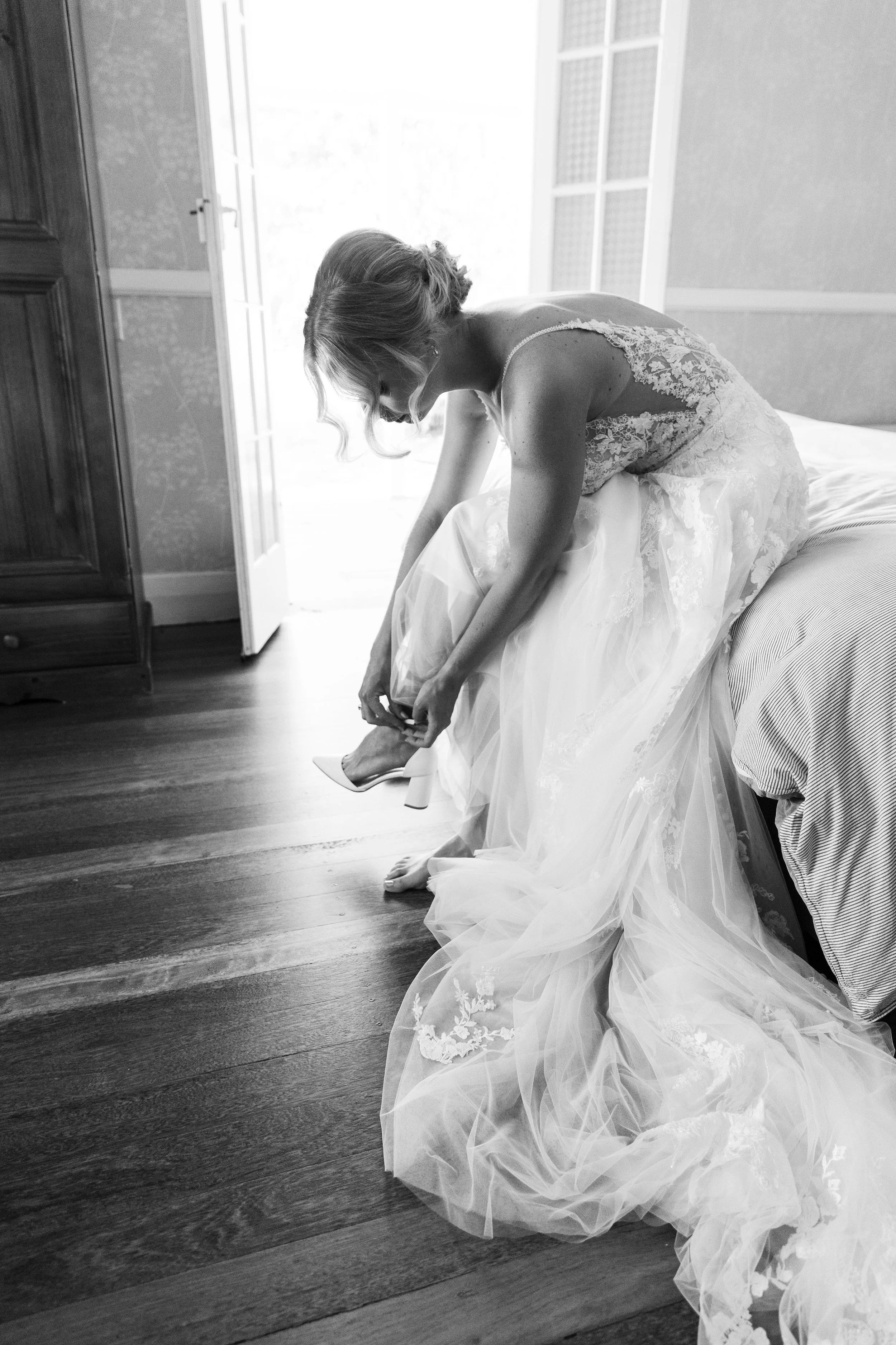 A bride in a lace wedding dress is sitting on a bed, bending forward to put on high-heeled shoes, near a window with natural light.