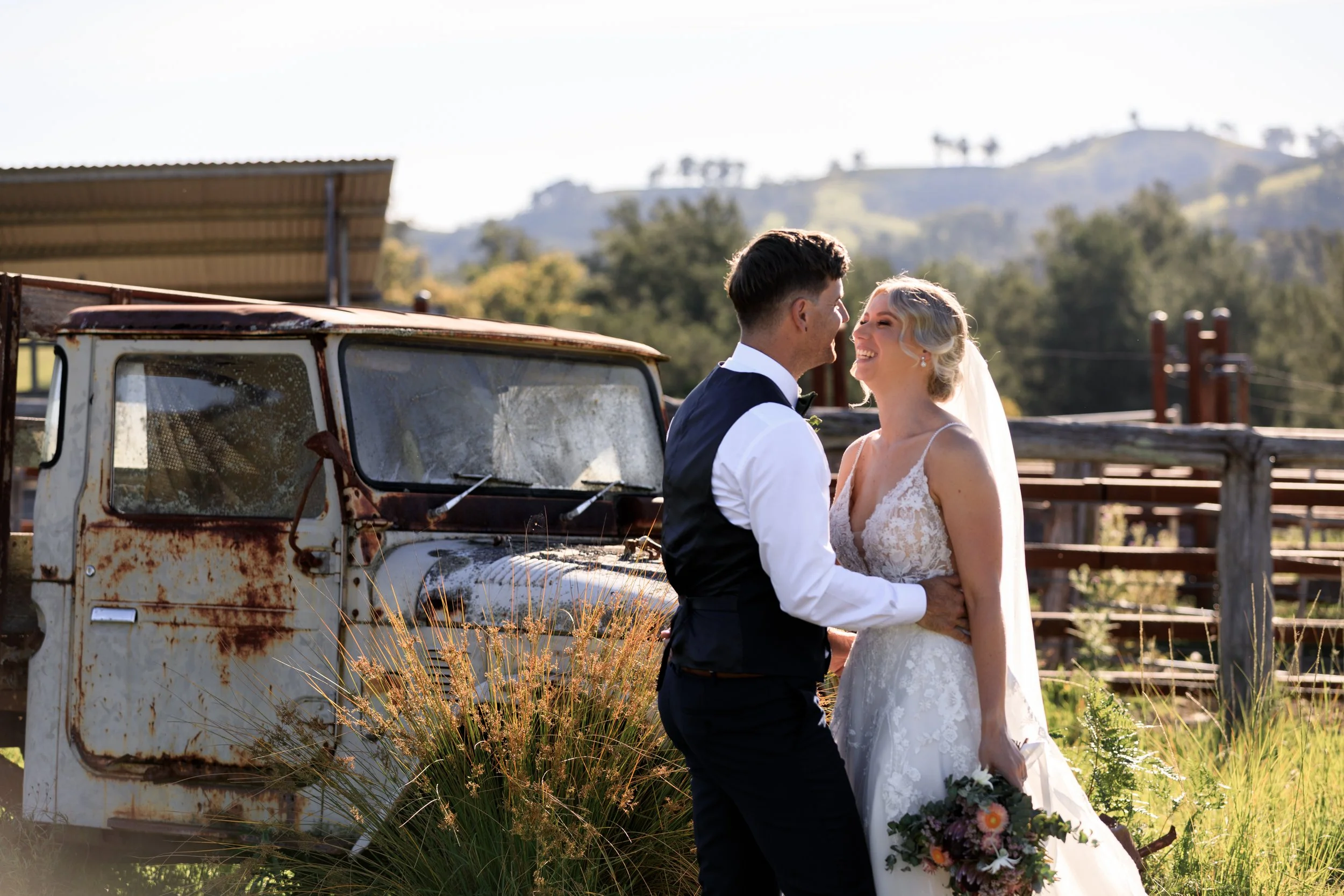 A bride and groom smiling and holding hands outdoors near an old rusty vehicle, with a rural landscape and hills in the background.
