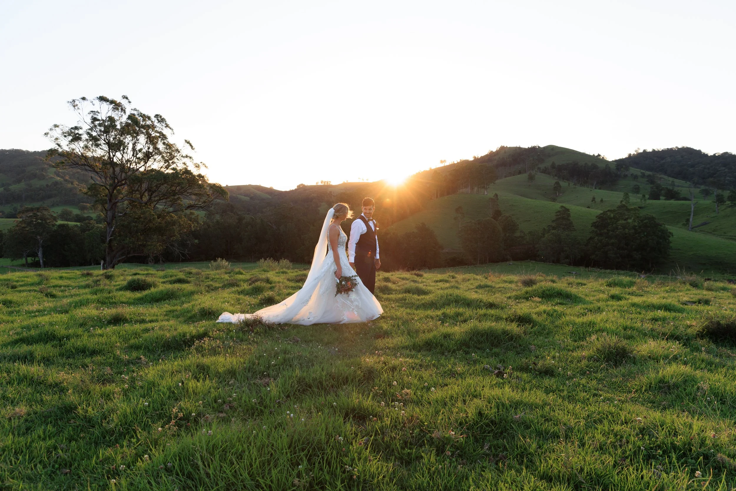 A bride and groom holding hands during their wedding in a green field at sunset with hills in the background.