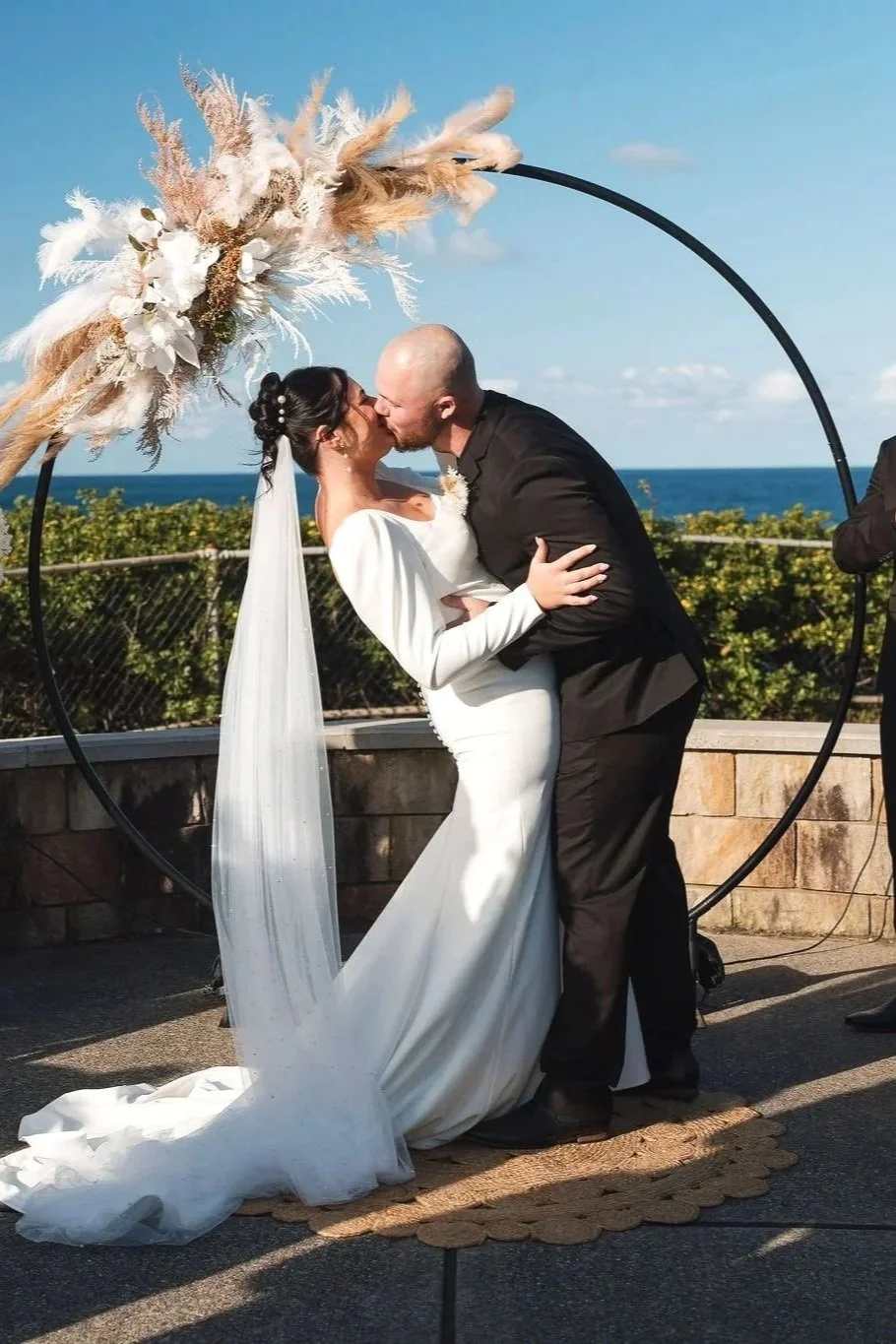 A bride and groom kissing under a circular wedding arch decorated with pampas grass and white flowers, with an ocean view in the background.